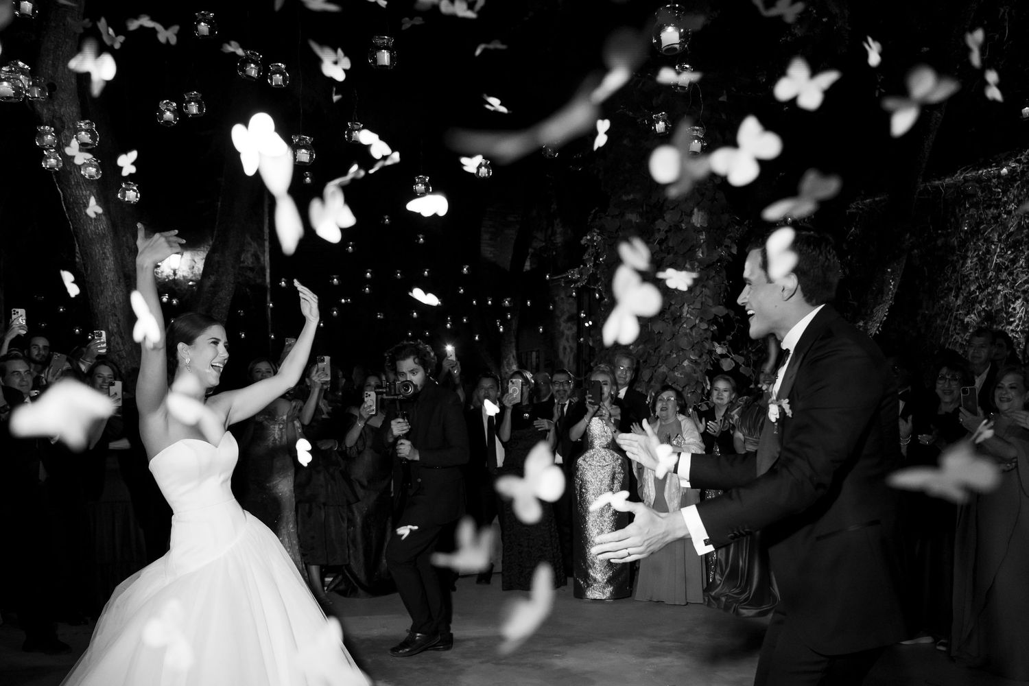 Black and white photo of bride and groom dancing as white butterflies or petals fill the air at elegant wedding reception.