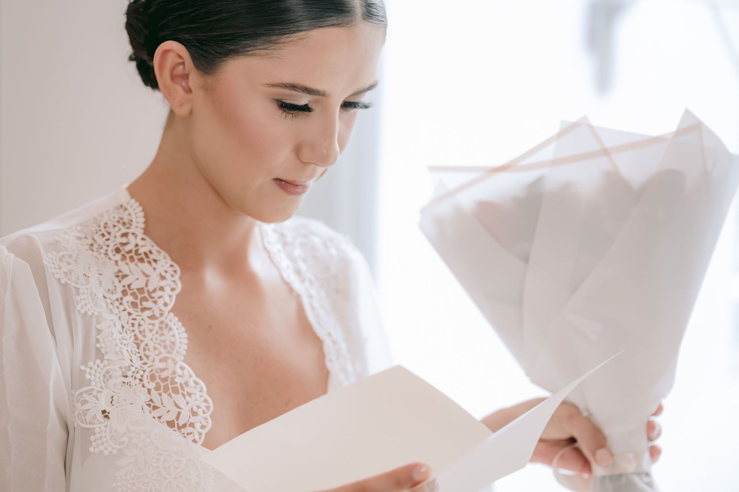 Elegant bride in white lace dress reading a card while holding a wrapped bouquet on her wedding day.