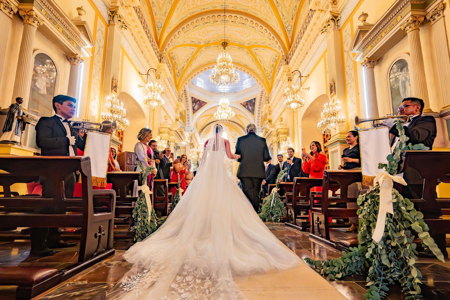 Bride walking down the aisle in an ornate golden church wearing a long white gown and veil during wedding ceremony.