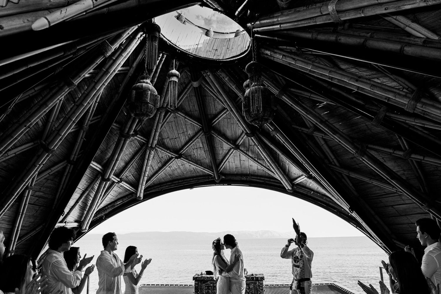Black and white wedding ceremony under bamboo arch with ocean view, couple kissing while guests applaud.