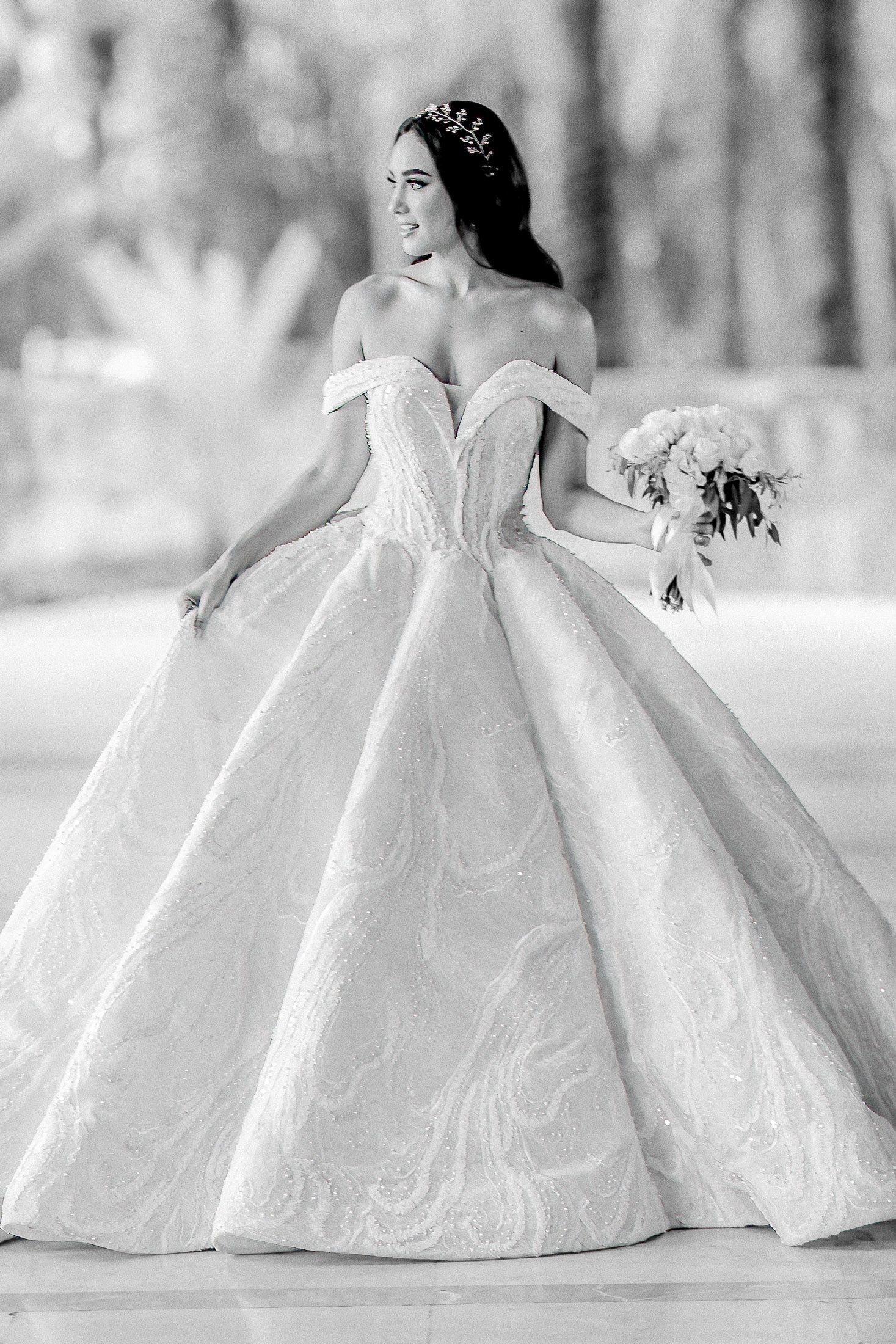 Elegant off-shoulder ball gown wedding dress with voluminous skirt photographed in black and white against outdoor backdrop.