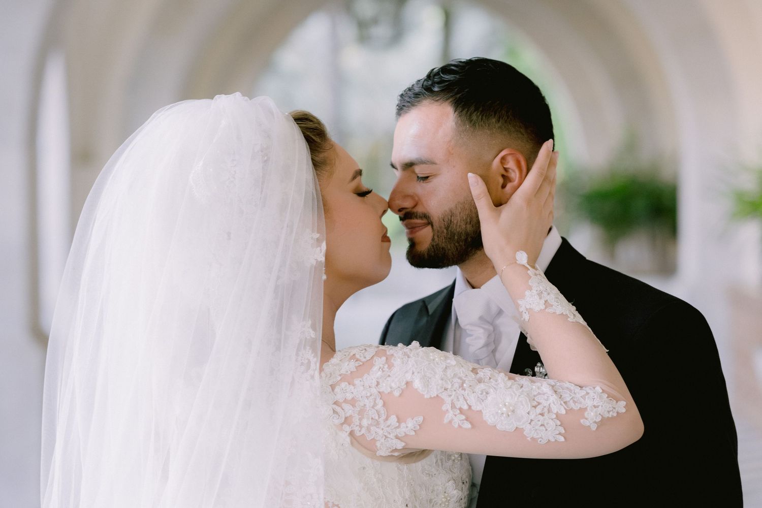 A romantic wedding couple shares a tender kiss in front of a blurred outdoor backdrop.
