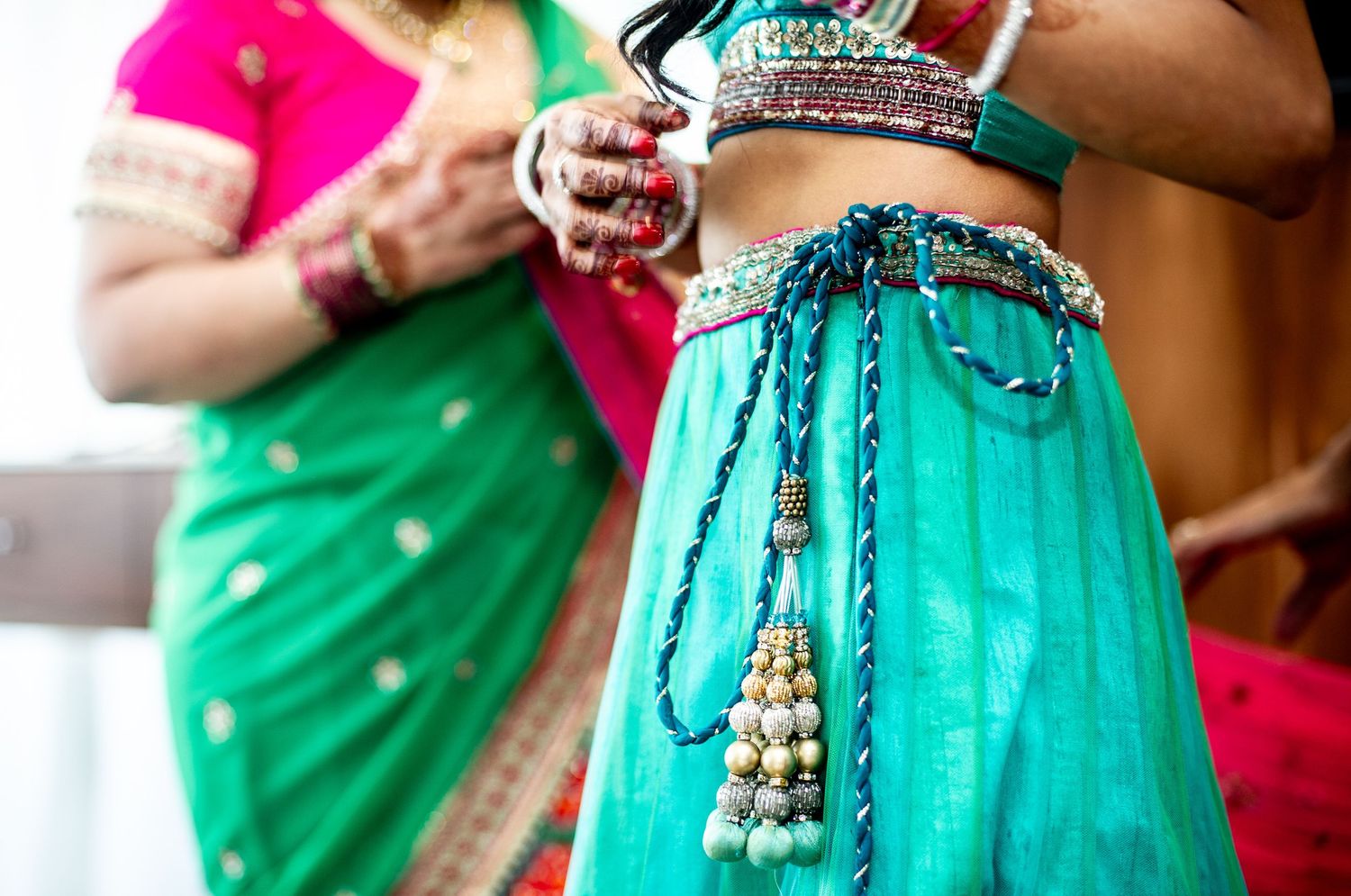 Two women in vibrant Indian traditional attire, featuring a teal lehenga with ornate jewelry and a green saree.