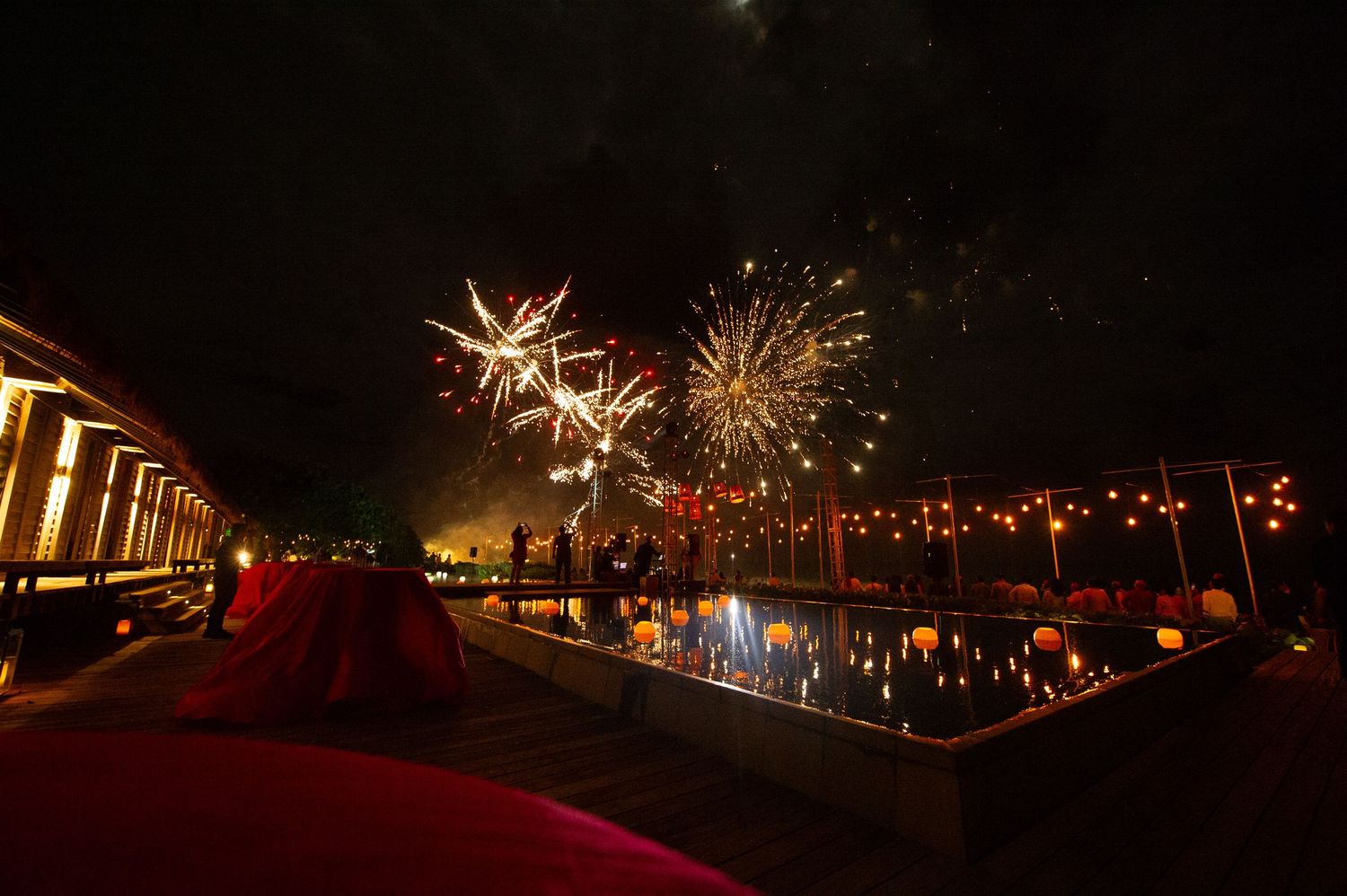 Colorful fireworks illuminate night sky over a festive poolside venue with warm lights and guests celebrating outdoors.