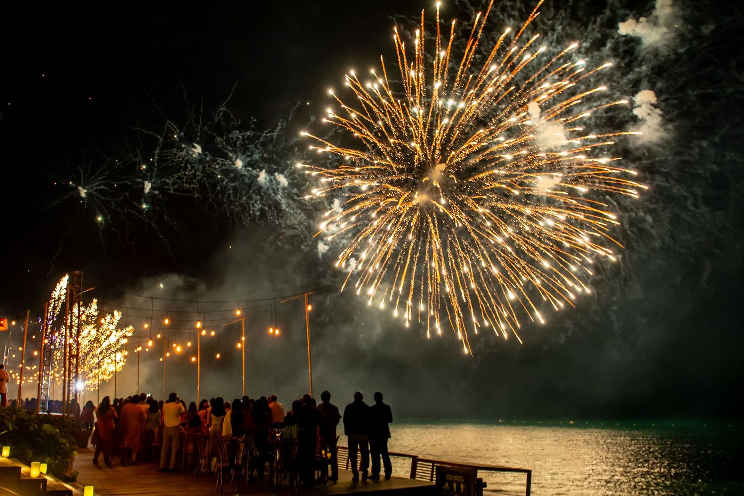 Crowd watching vibrant golden fireworks burst over a waterfront at night during a festive celebration.