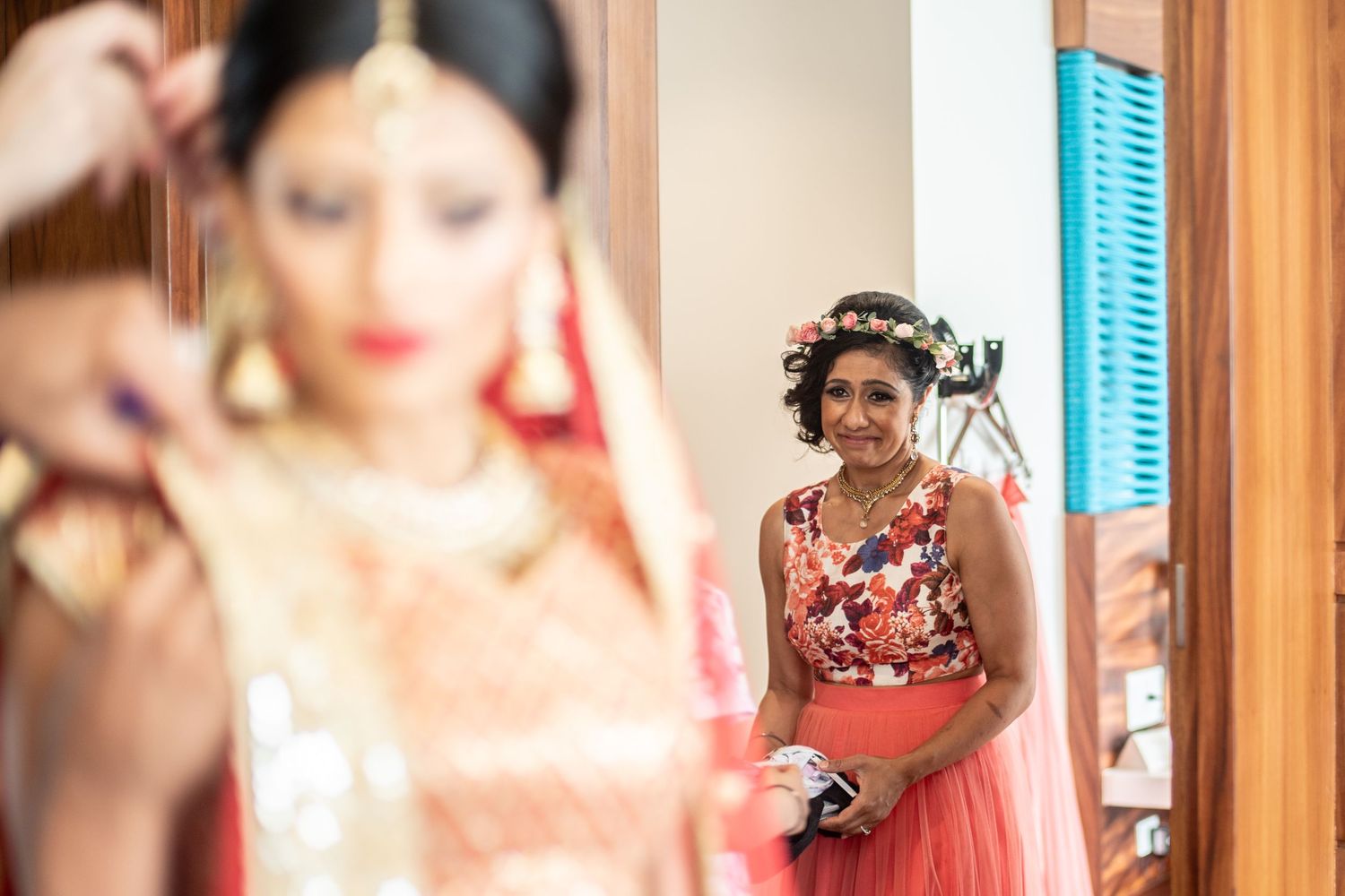 Indian bride in traditional red and gold bridal attire being prepared, with a smiling woman in floral dress watching in background.