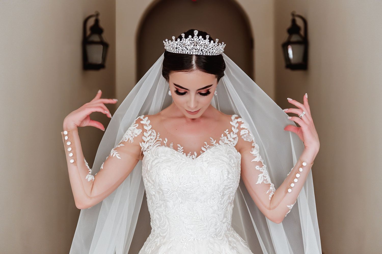 Elegant bride in lace wedding gown and crystal tiara poses with flowing veil in archway.