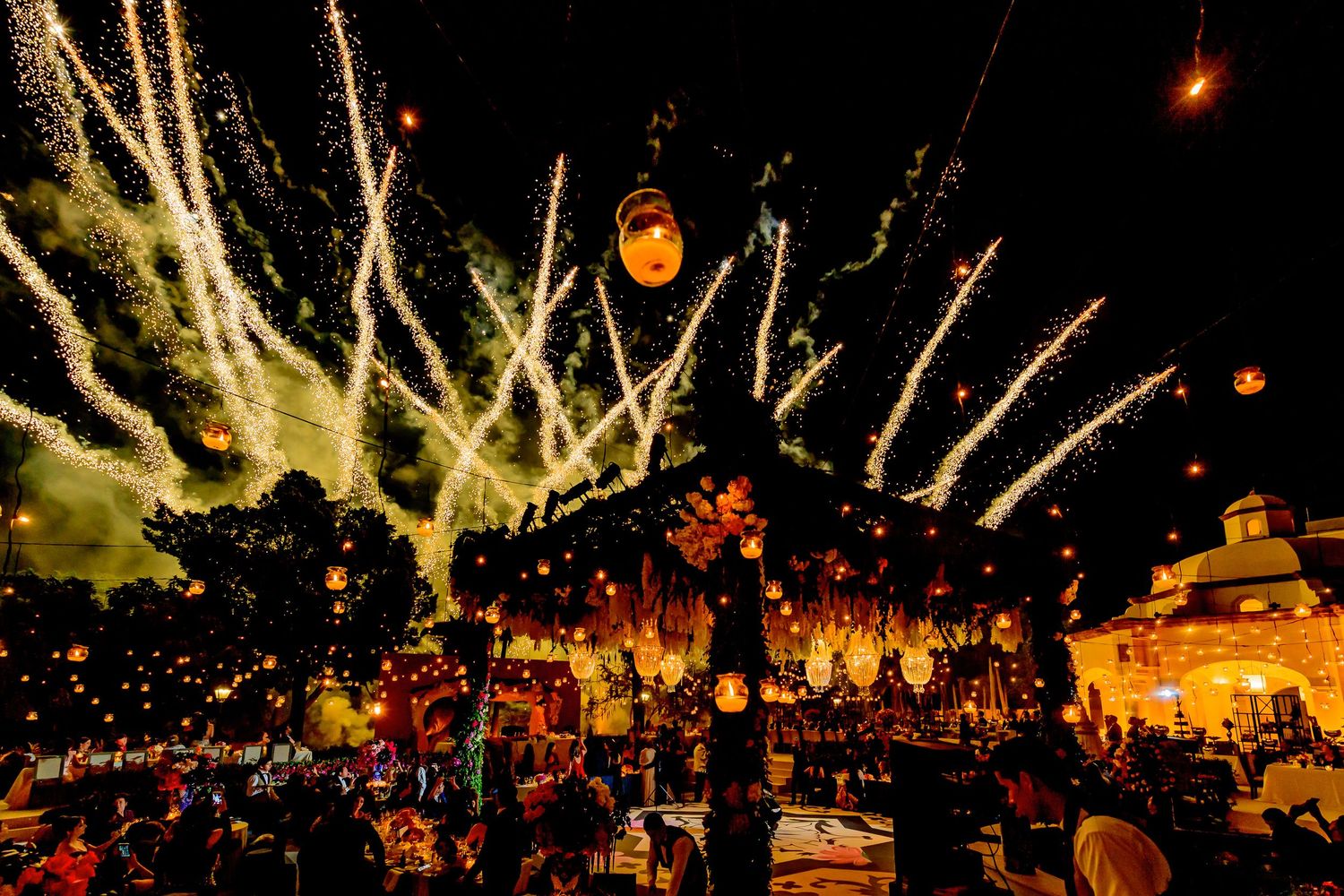 Glowing lanterns illuminate a nighttime outdoor celebration with architectural elements in the background.
