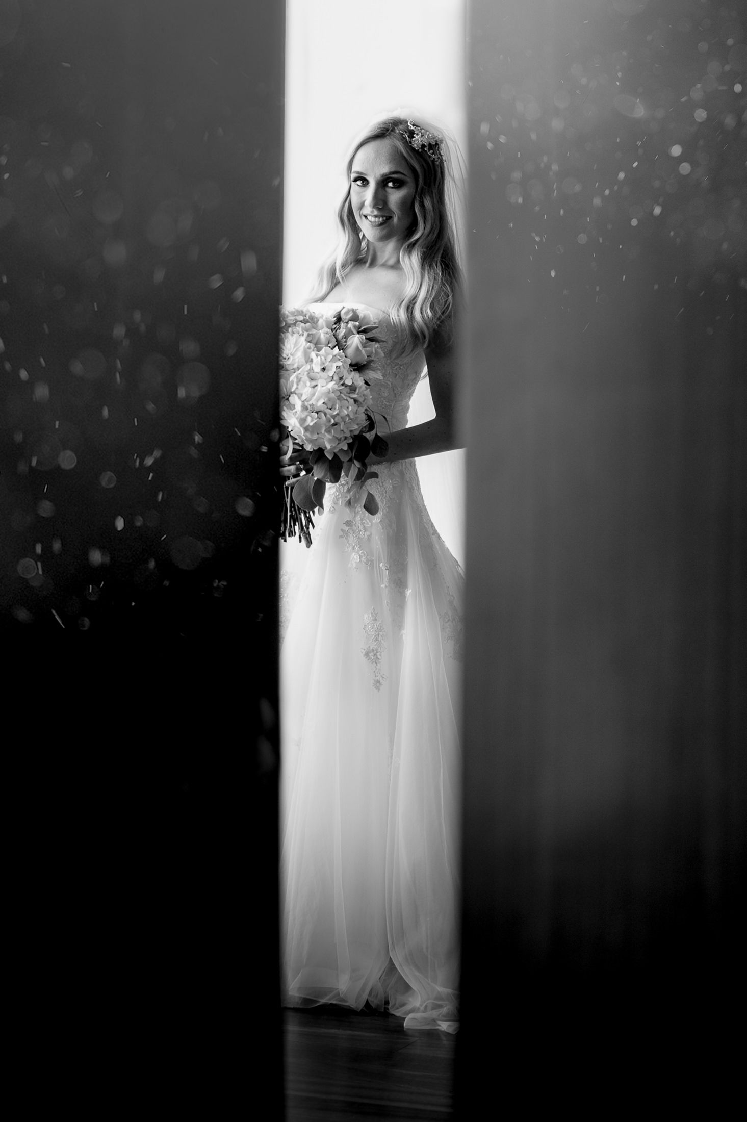 Ethereal black and white photo of a bride holding bouquet while standing in a beam of light.