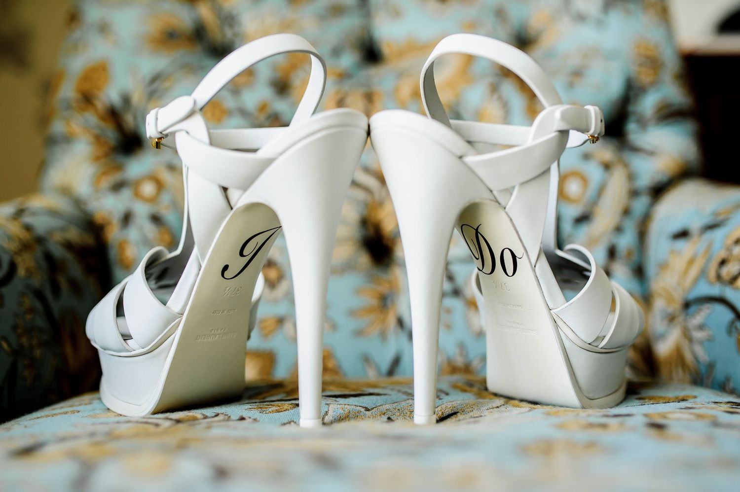 White wedding heels with 'I Do' written on soles displayed on blue floral chair.