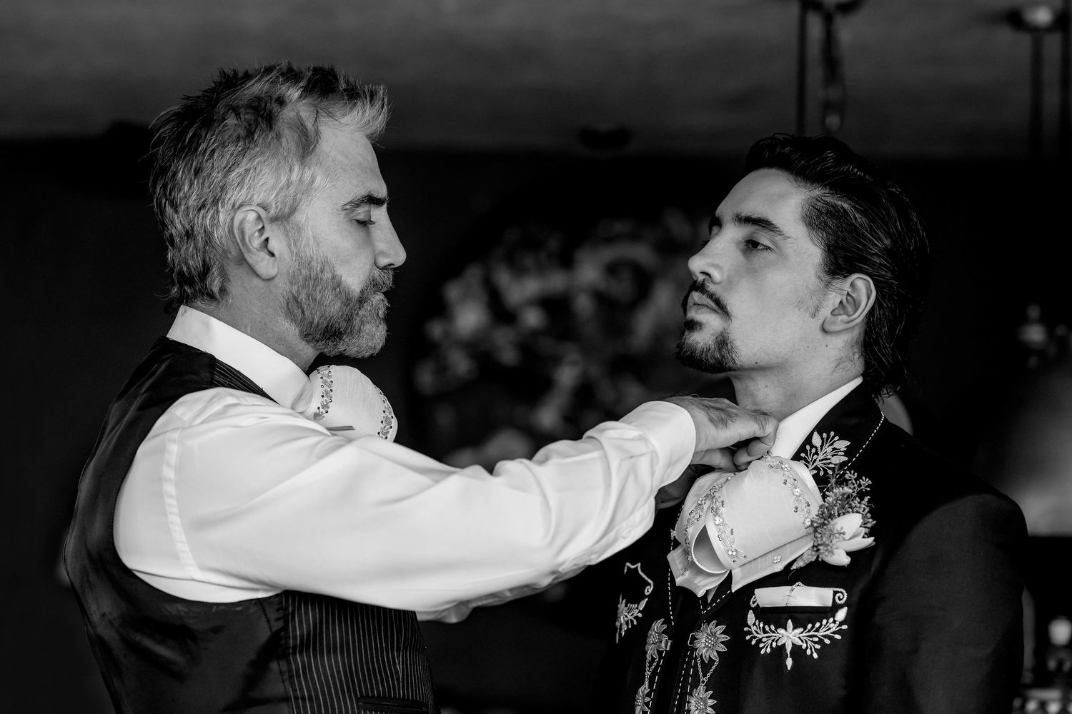 Black and white photo of two men in formal attire, one adjusting the other's boutonniere at a wedding.