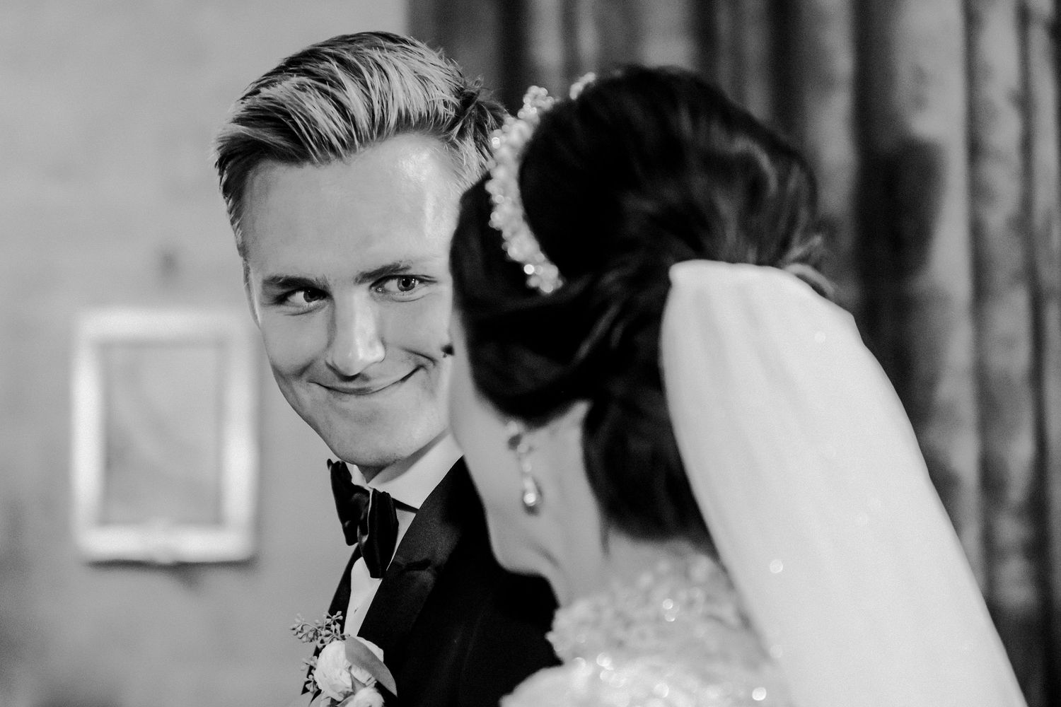 Black and white wedding photo of a smiling groom in a tuxedo looking at his bride in a pearl hair accessory.