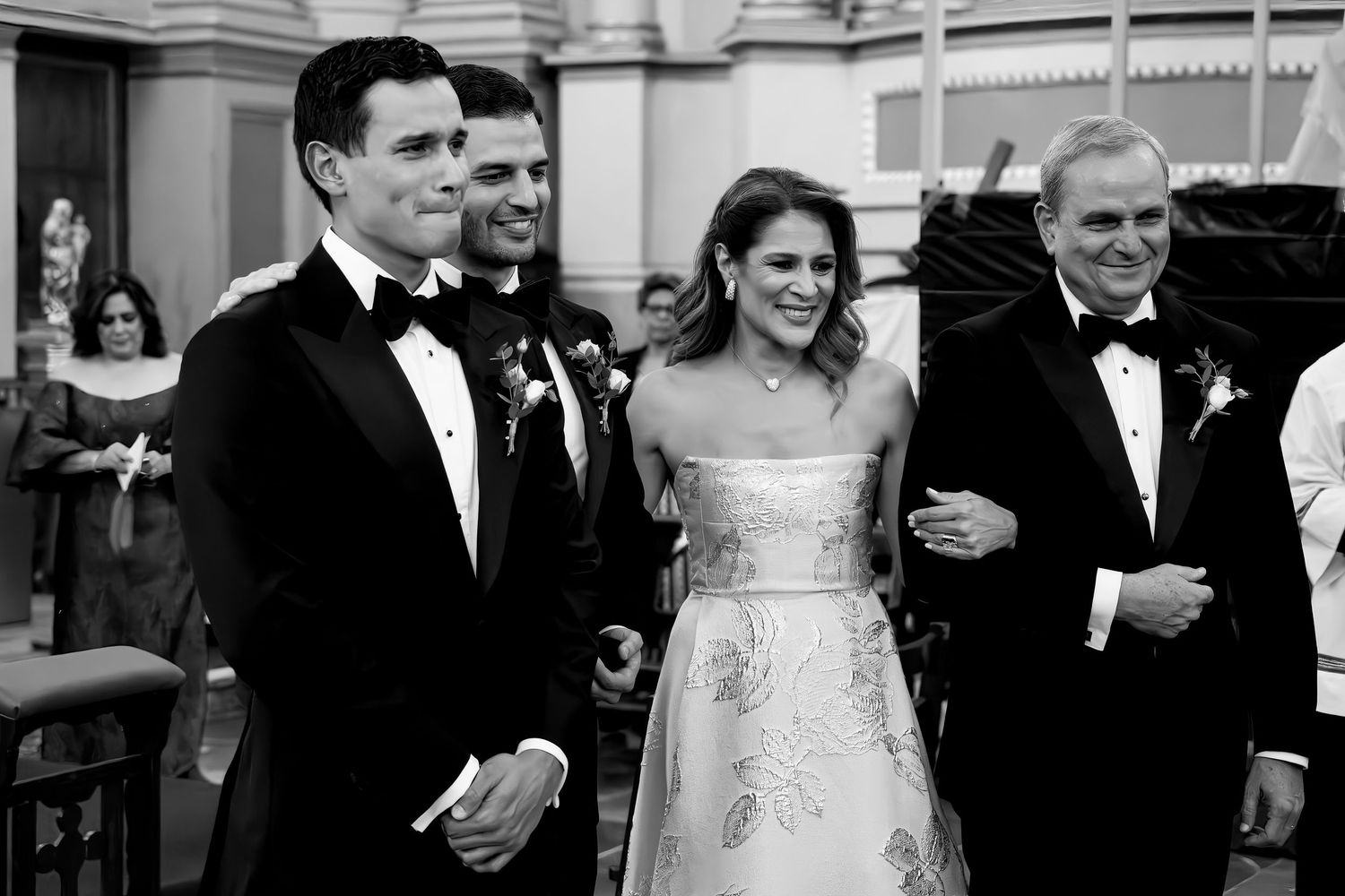 Black and white wedding photo of bride in strapless gown smiling with groom and guests in formal attire.