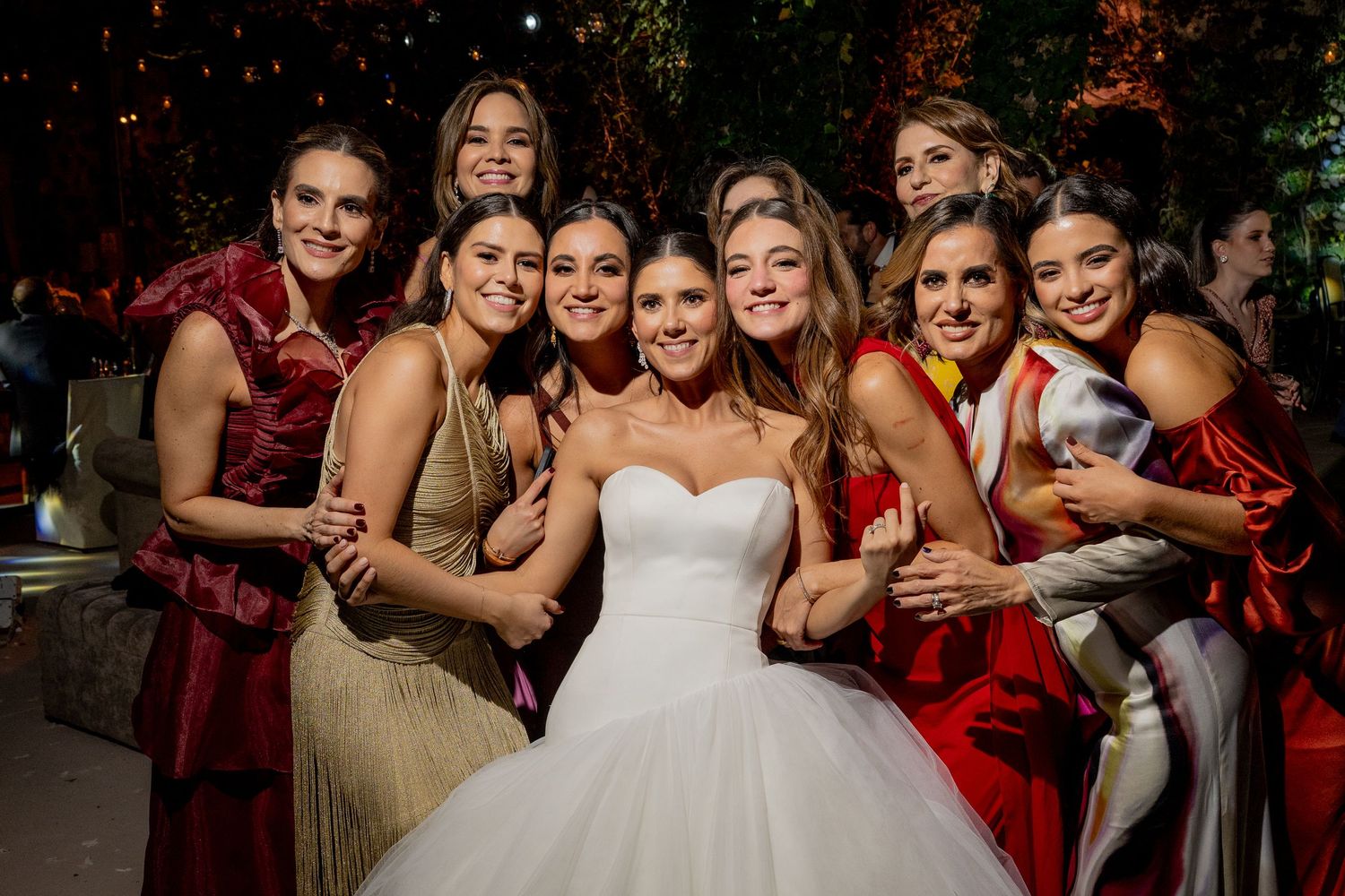 Bride in white ball gown poses with bridesmaids in red and gold dresses at elegant outdoor wedding reception.