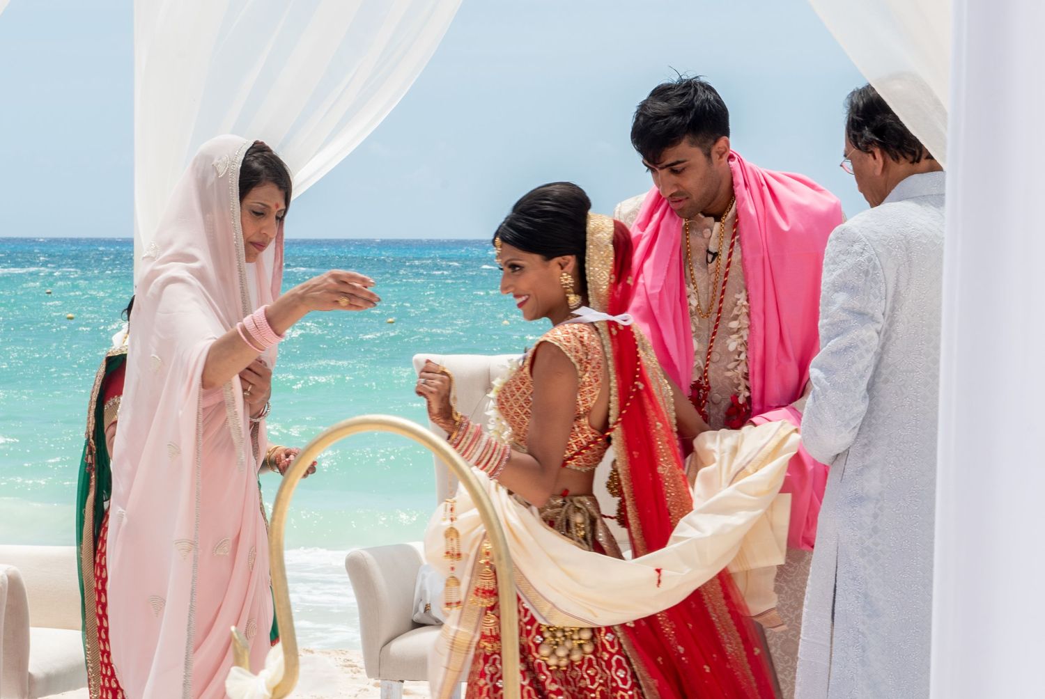 Indian wedding ceremony on a beach with bride in red lehenga, groom in pink sherwani, and family members assisting.