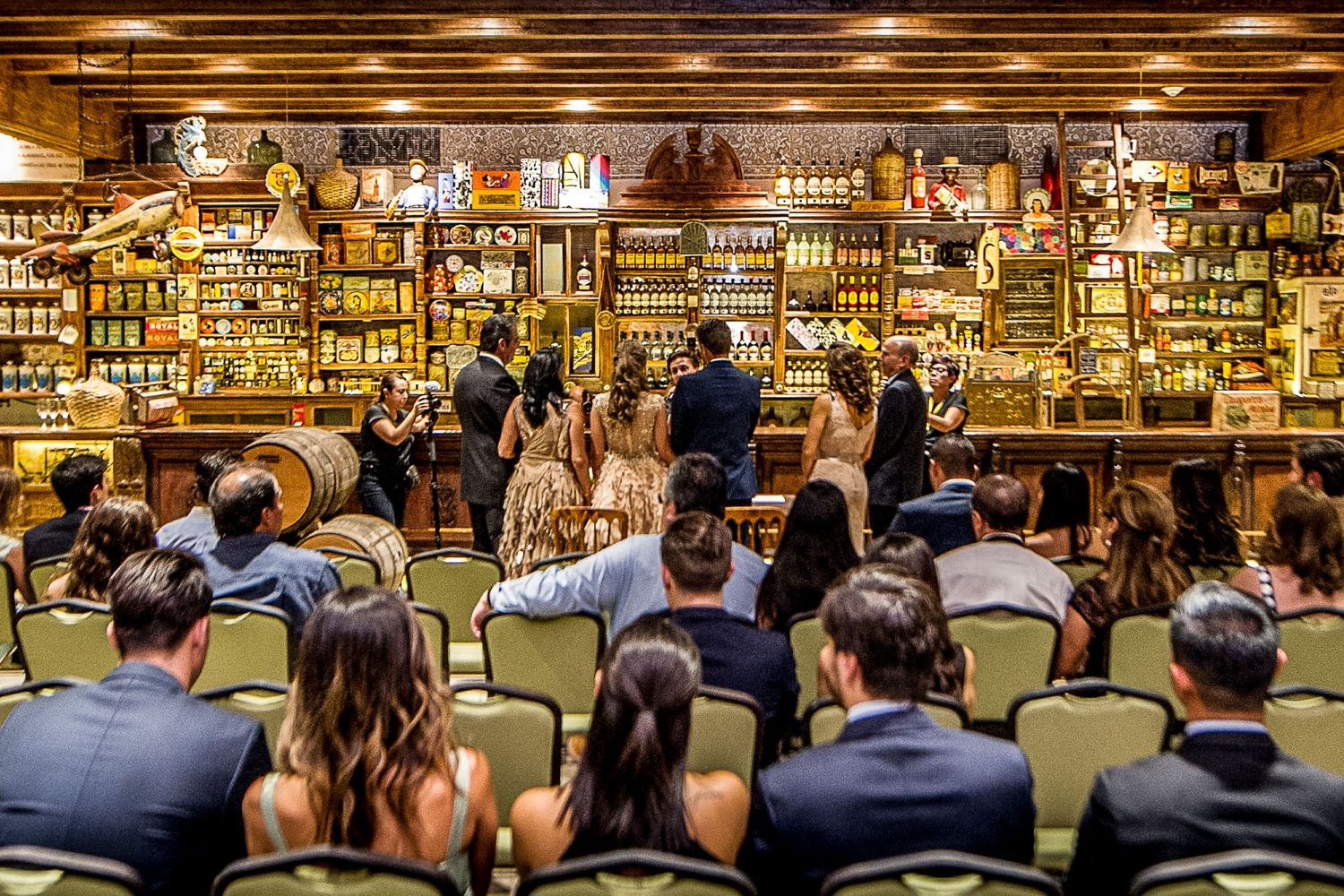 Guests seated at a rustic bar venue during a wedding ceremony with wooden shelves lined with bottles.