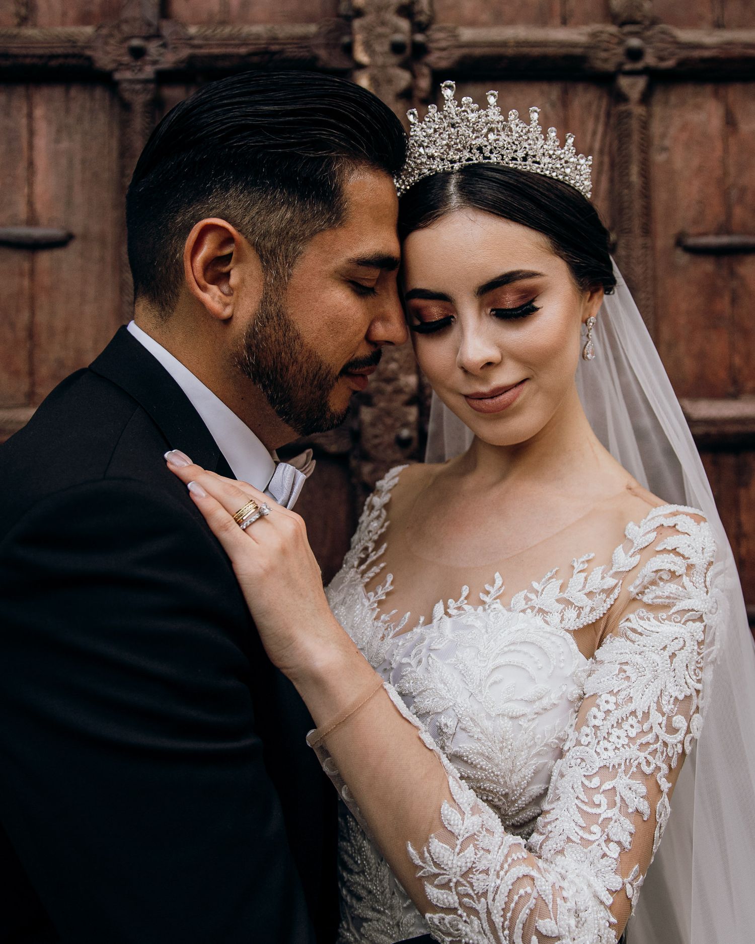 A bride in lace gown and crystal tiara shares an intimate moment with her groom in a black tuxedo near ornate wooden doors.