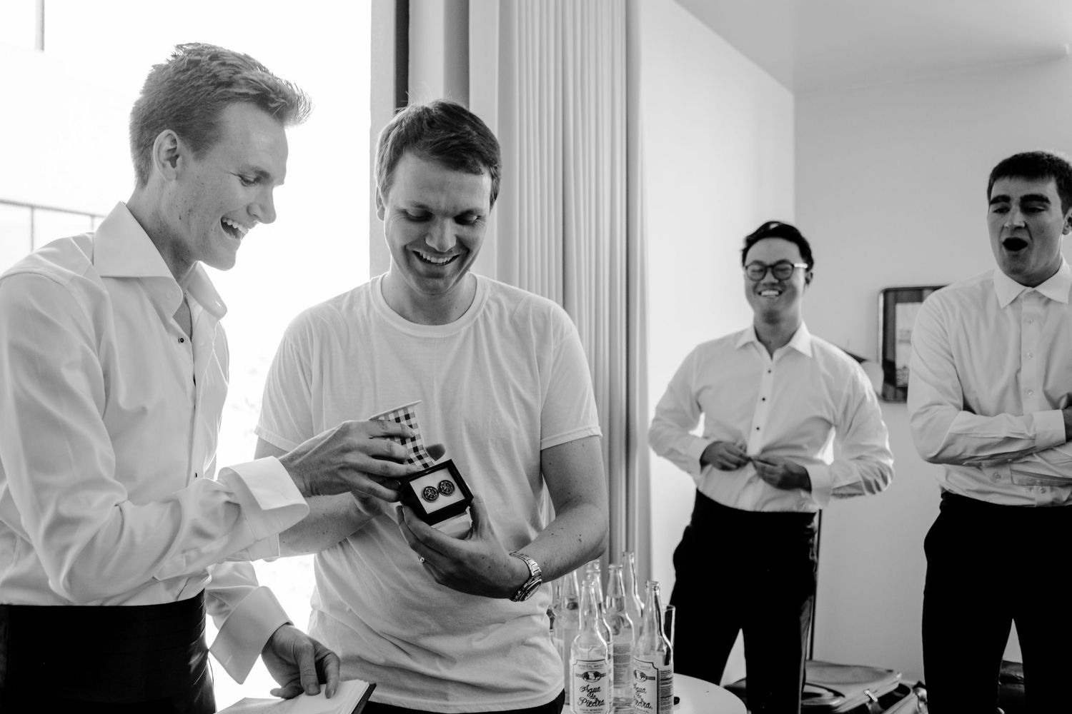 Black and white photo of men laughing and opening a gift at a wedding preparation gathering.
