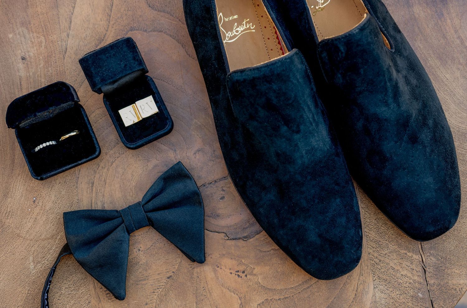 Navy velvet loafers, black bow tie, gold cufflinks, and a ring box arranged on a wooden surface for a wedding.
