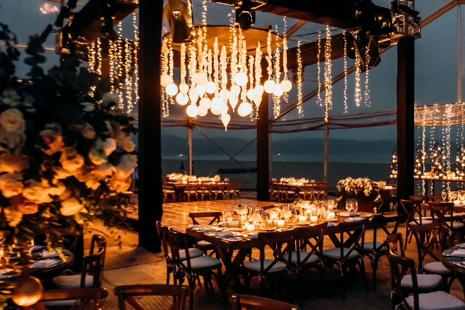 Sunset wedding reception overlooking the water with hanging crystal chandeliers and string lights.