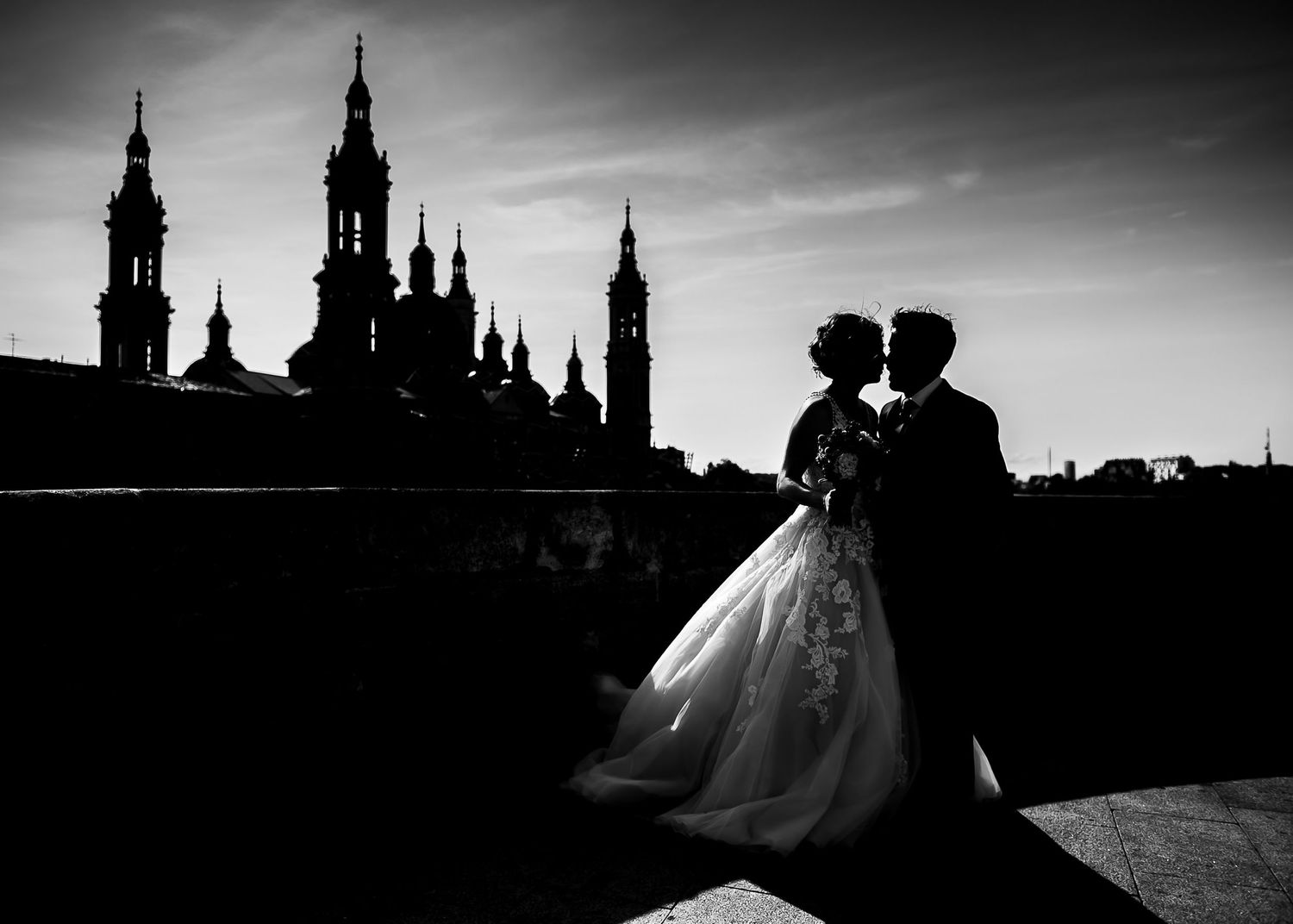 Silhouette of wedding couple against dramatic sky with cathedral spires in background.