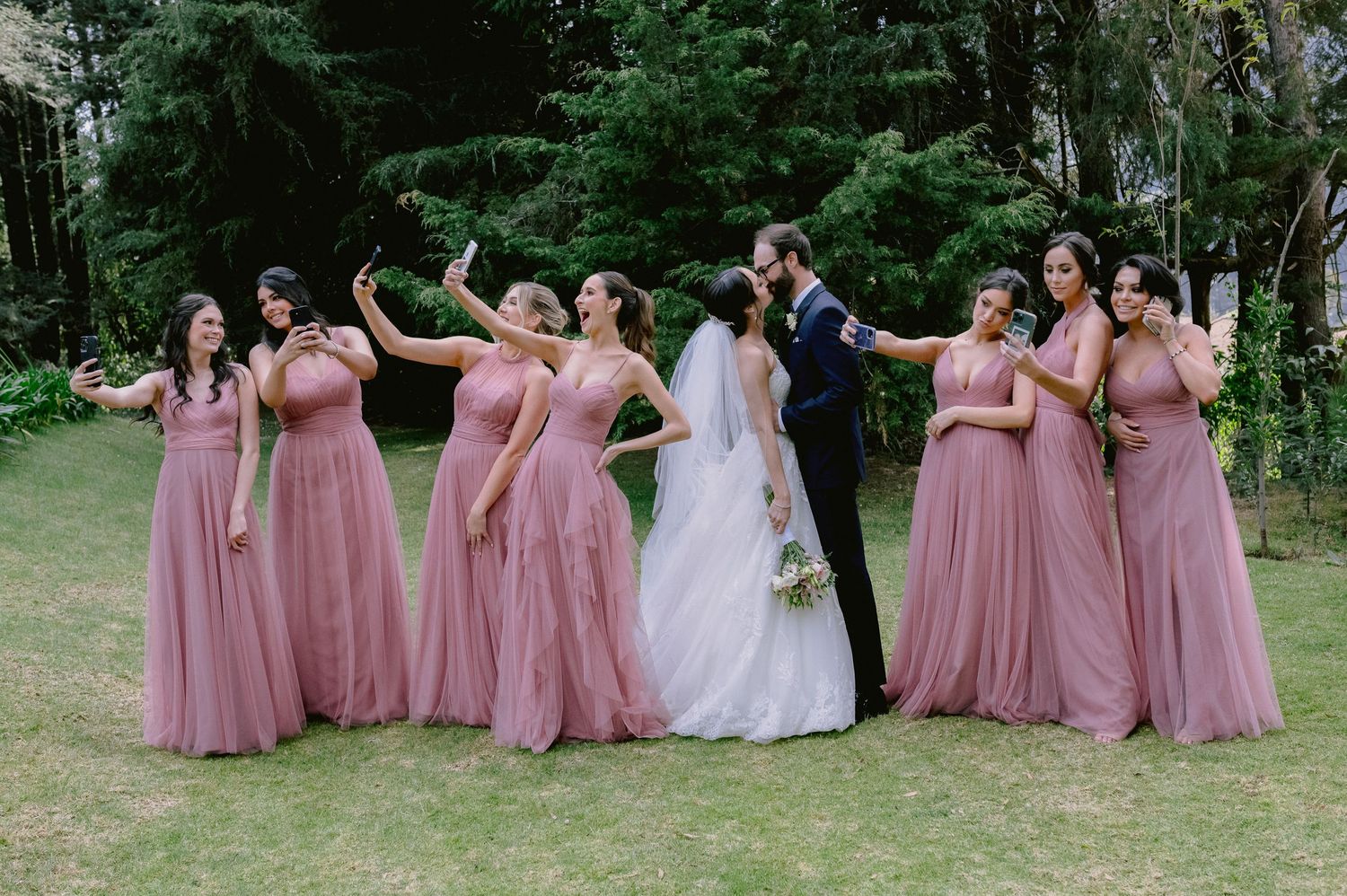 Wedding party in dusty rose bridesmaid dresses poses together outdoors in a garden setting.