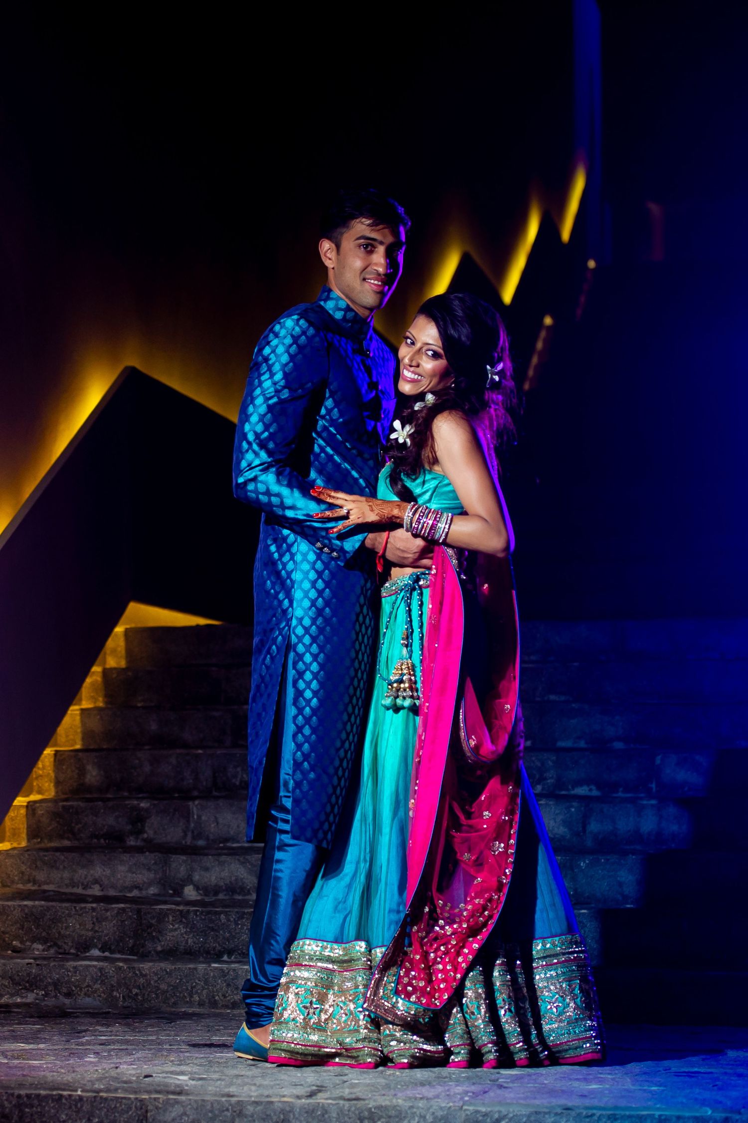 Indian couple in traditional attire, man in blue sherwani, woman in teal and pink lehenga, posing by stairs.