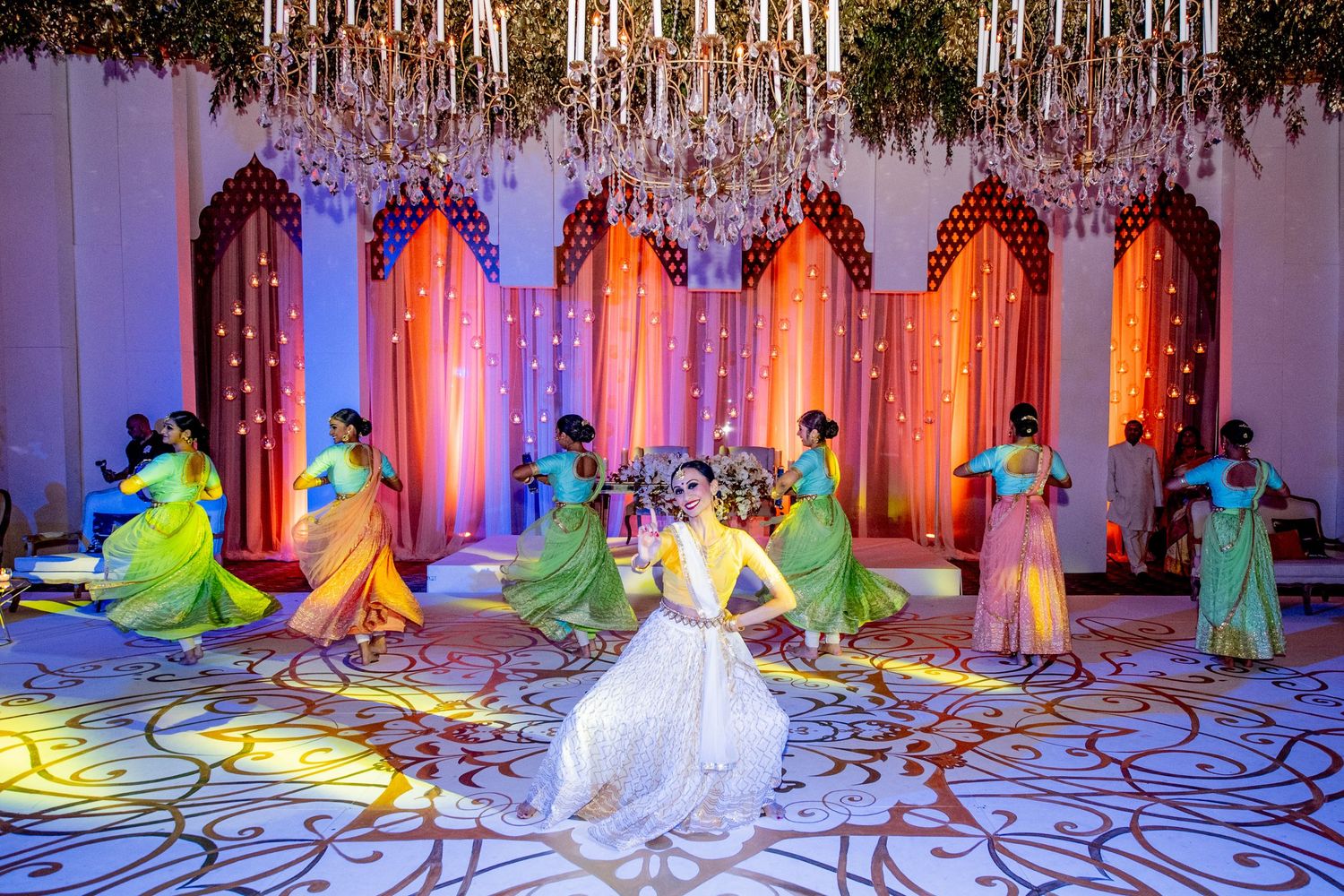 Bride in white gown surrounded by dancers in colorful sarees performing at an elegant Indian wedding celebration.