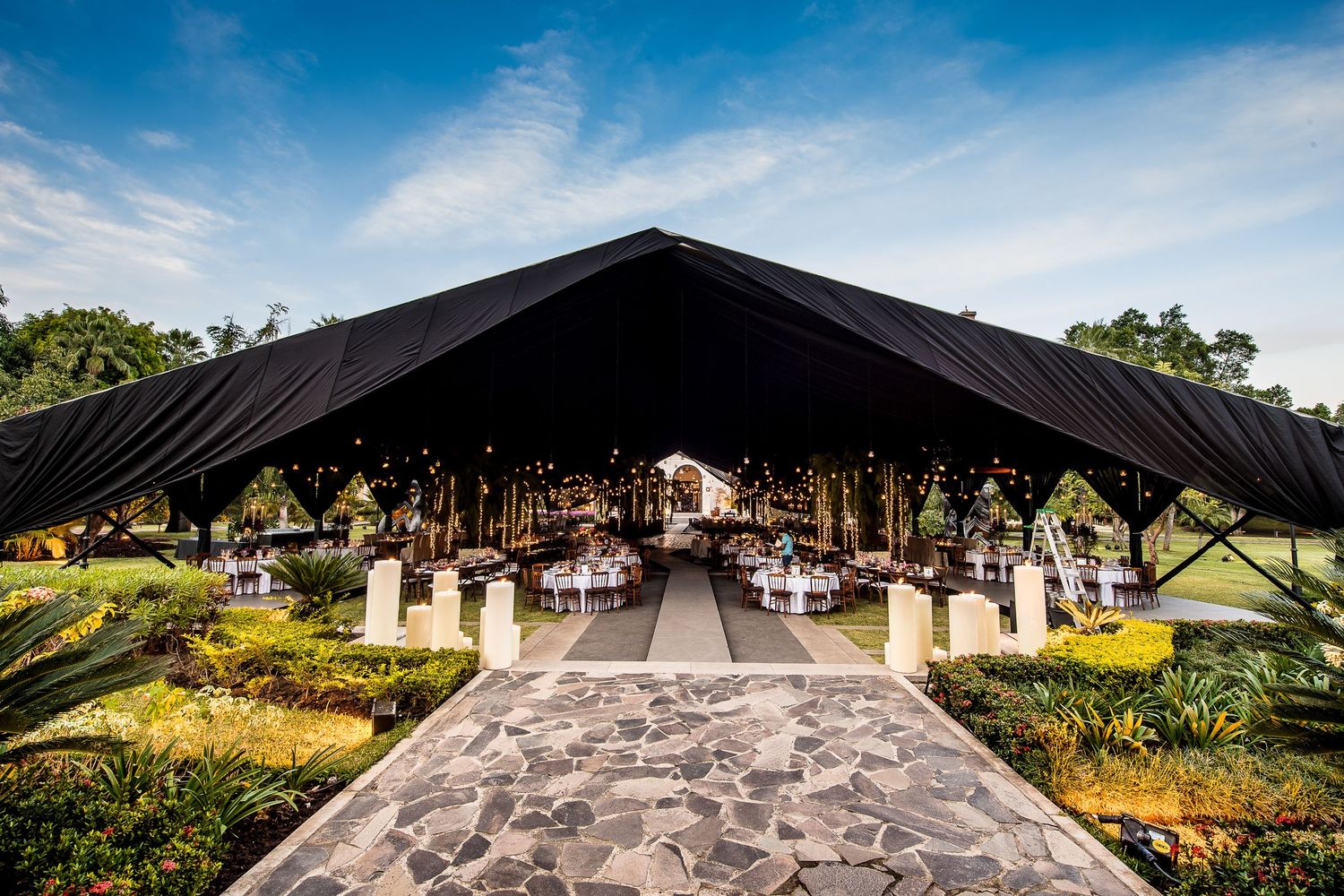 Elegant outdoor event venue with black tent canopy, decorated tables, string lights, and stone pathway surrounded by lush greenery.