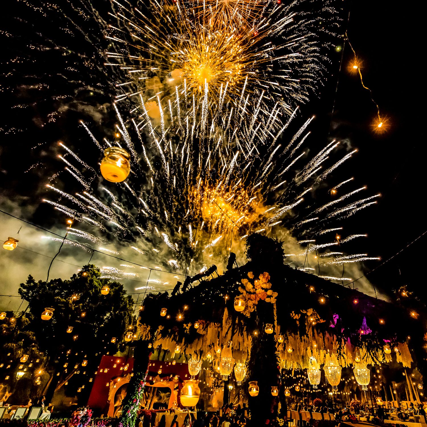 Golden fireworks display illuminates the night sky over an outdoor celebration with hanging lanterns.