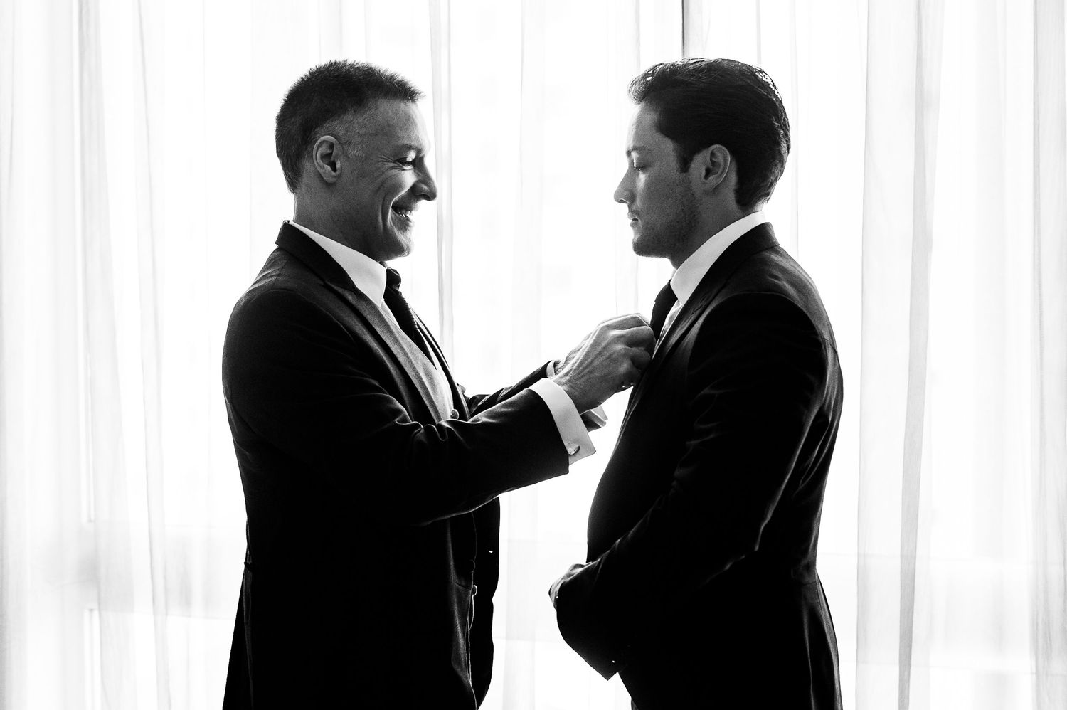 Two men in formal suits adjusting their ties before a wedding ceremony.