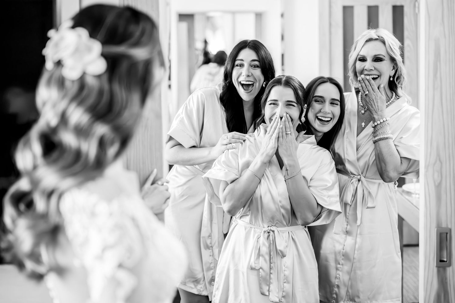 Bridesmaids in white robes react with joy and excitement as bride reveals her wedding look in black and white photo.