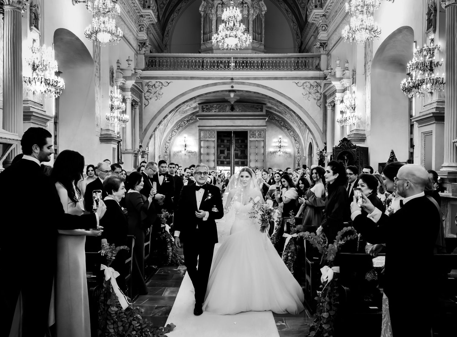 Black and white photo of bride and groom walking down the aisle of an elegant chandelier-lit church.