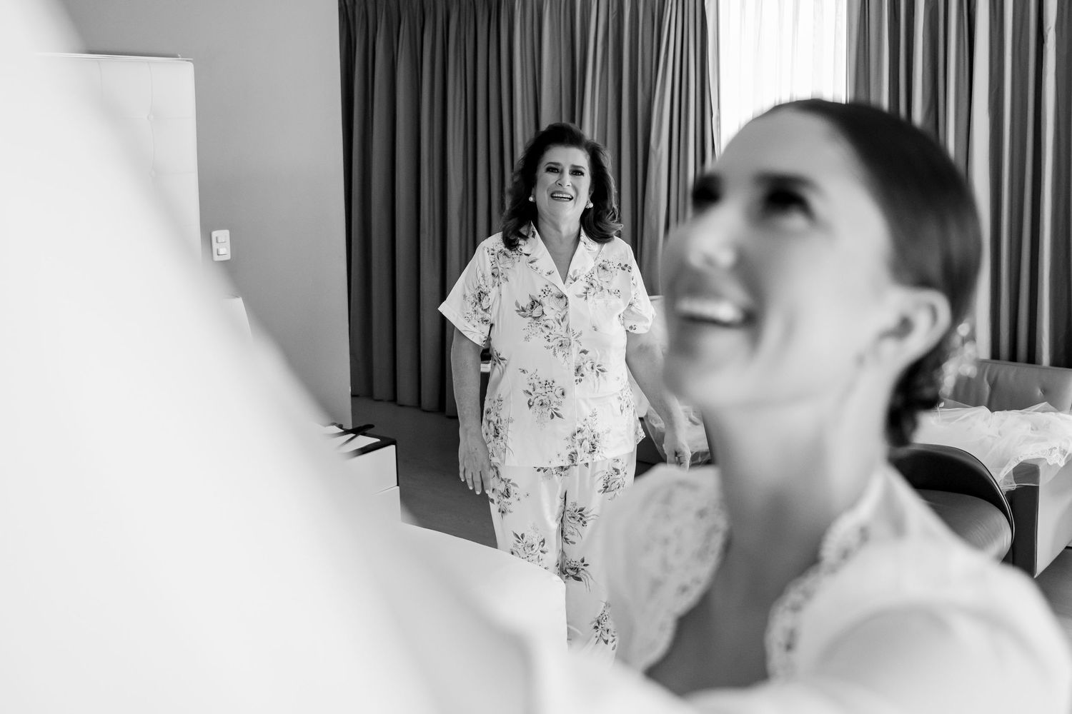 Black and white photo of a bride laughing joyfully while a woman in floral pajamas smiles warmly in the background.