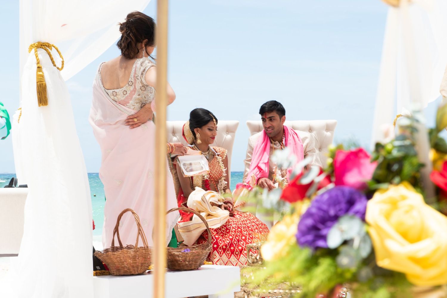 Colorful Indian wedding ceremony on a beach with bride and groom in traditional attire surrounded by vibrant floral decorations.