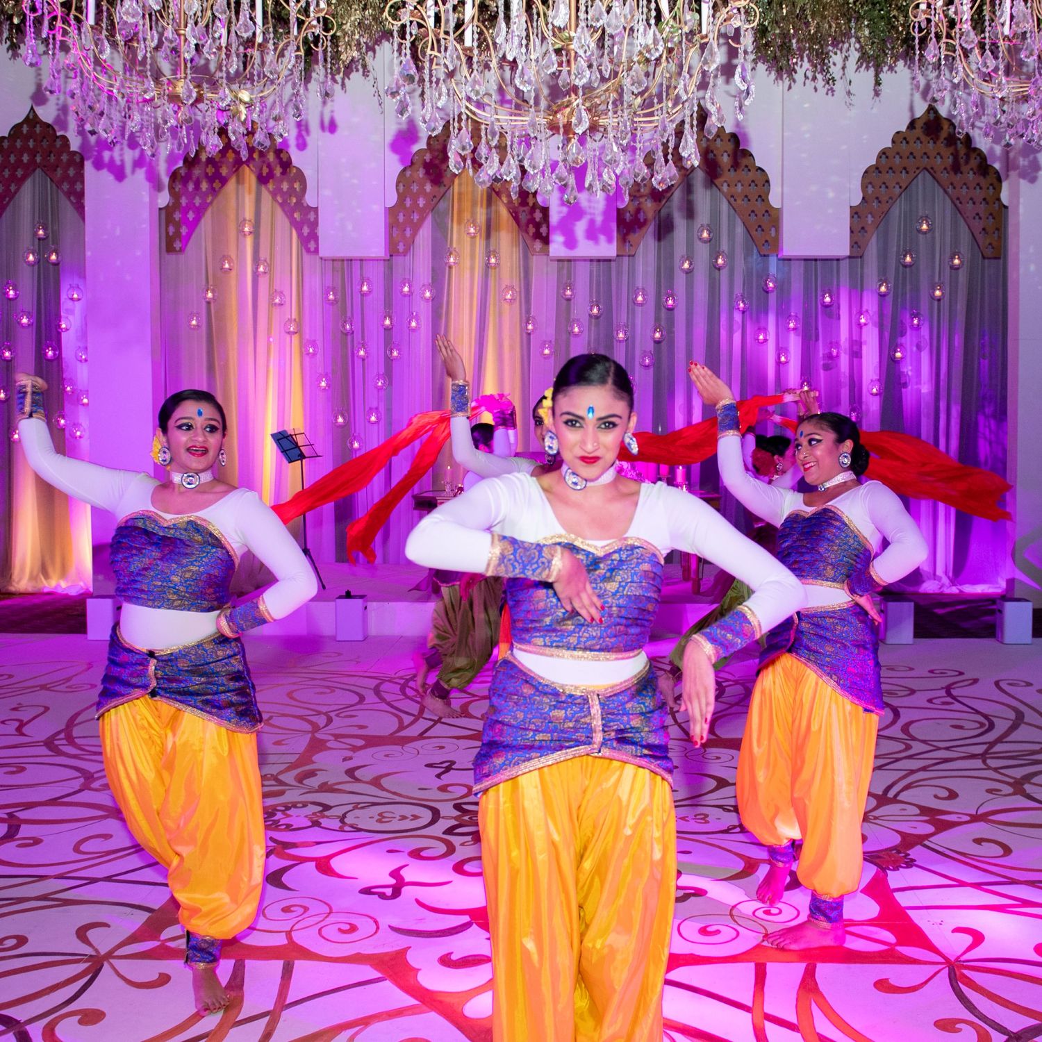 Three dancers in purple and gold costumes perform Bollywood dance at an elegant decorated Indian wedding reception.