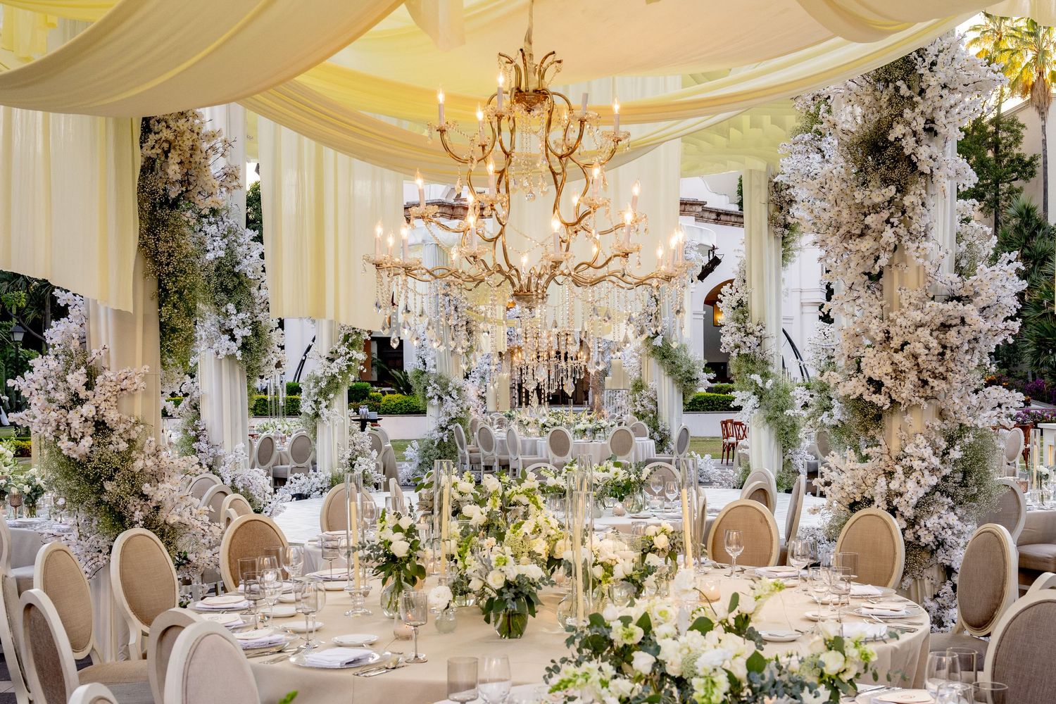 Luxurious wedding reception setup with crystal chandeliers, white draping, and lush floral arrangements.