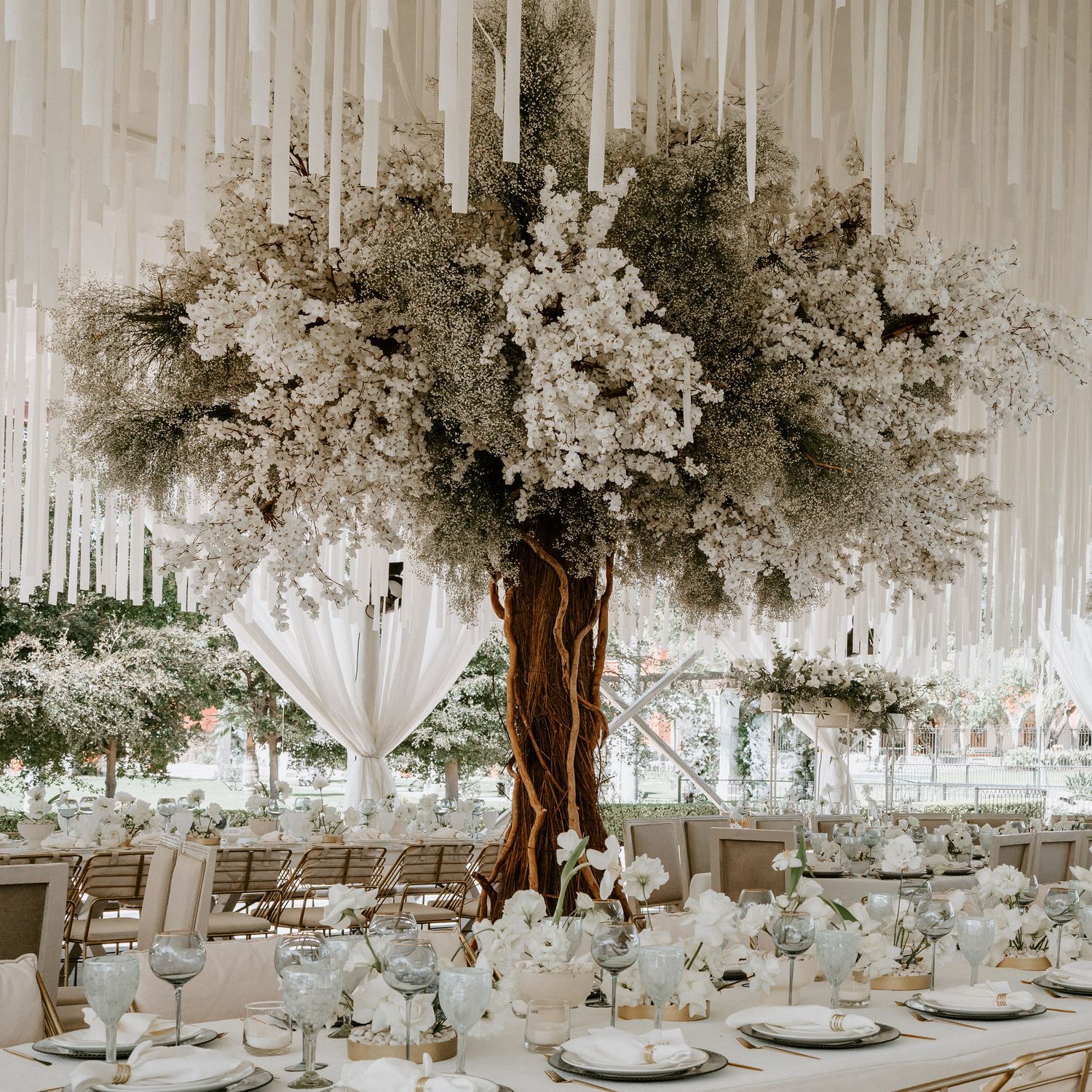 White-themed wedding reception space with artificial trees and elegant table settings creating a dreamy atmosphere.