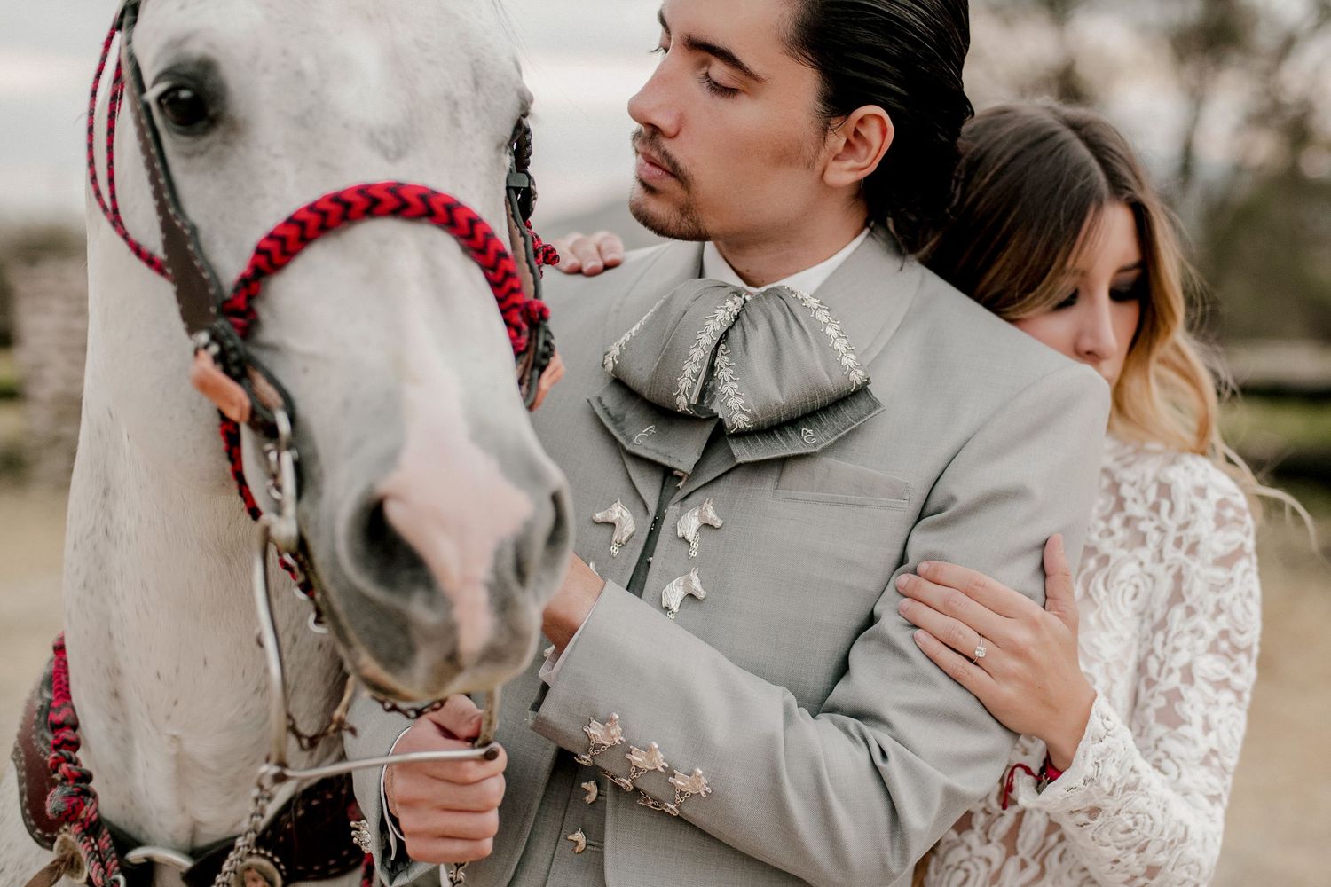Sweet moment between couple and white horse with red bridle in outdoor setting.