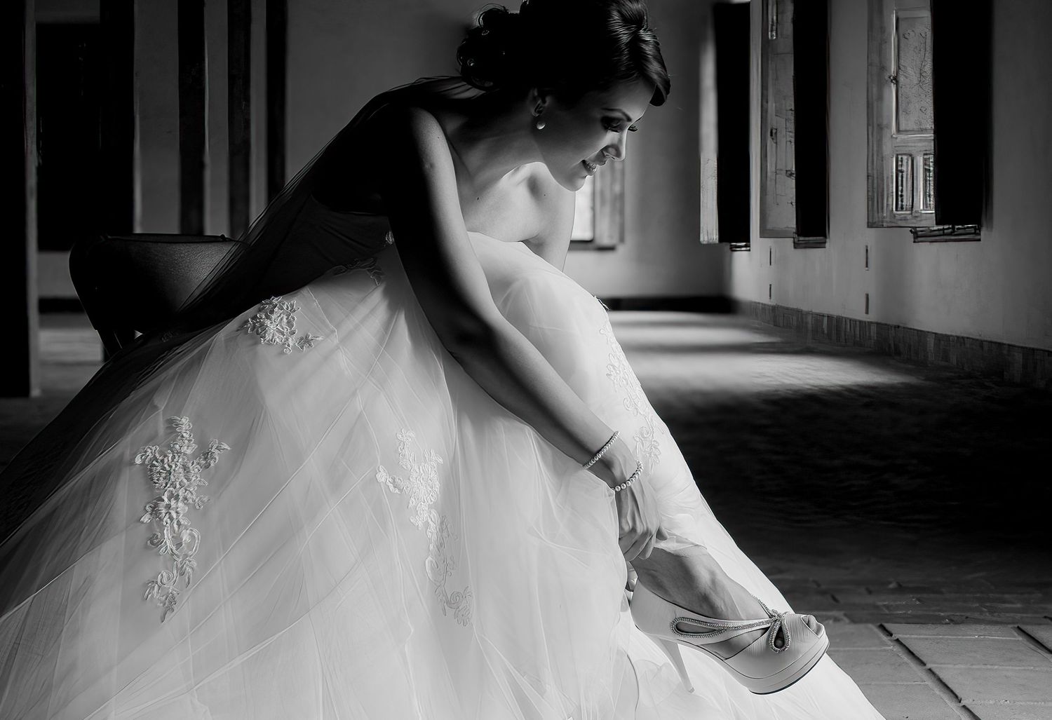 Black and white image of a bride in a tulle gown adjusting her shoe in an elegant setting.