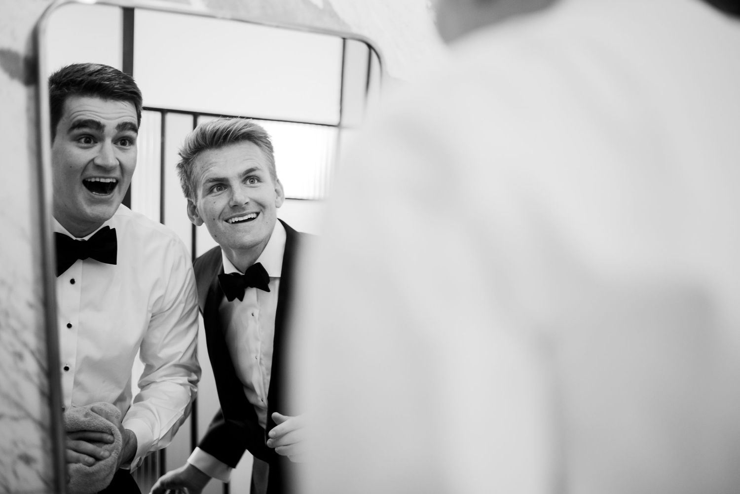 Two groomsmen in bow ties laughing together near mirror in black and white wedding photo.
