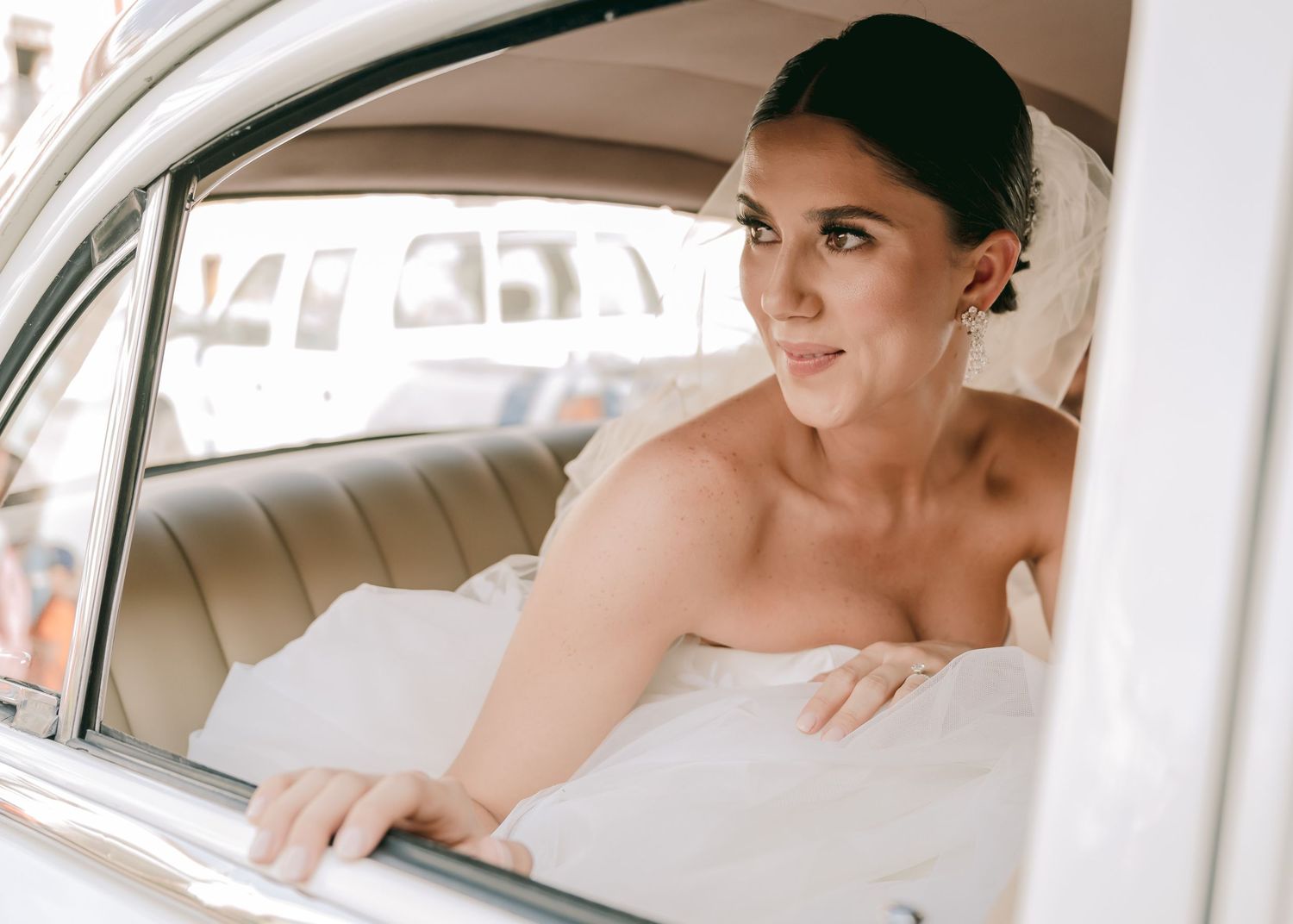 Bride in a white wedding dress sitting in a vintage car, looking elegantly over her shoulder.