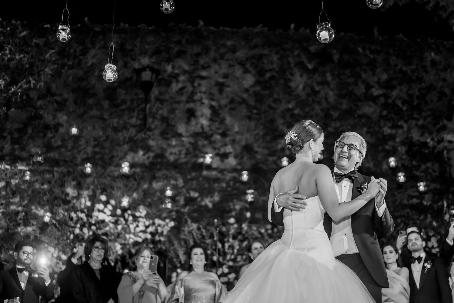 Black and white photo of bride and groom sharing first dance under string lights with guests watching at elegant wedding.