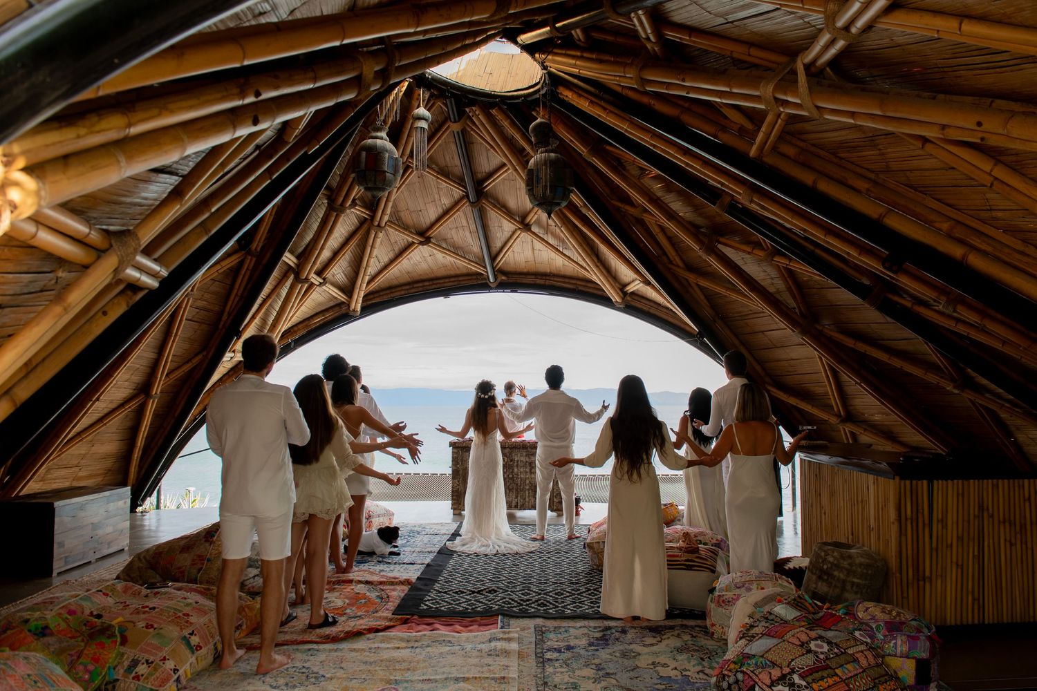 Wedding ceremony inside a bamboo dome structure with ocean views, bridal party in white holding hands.
