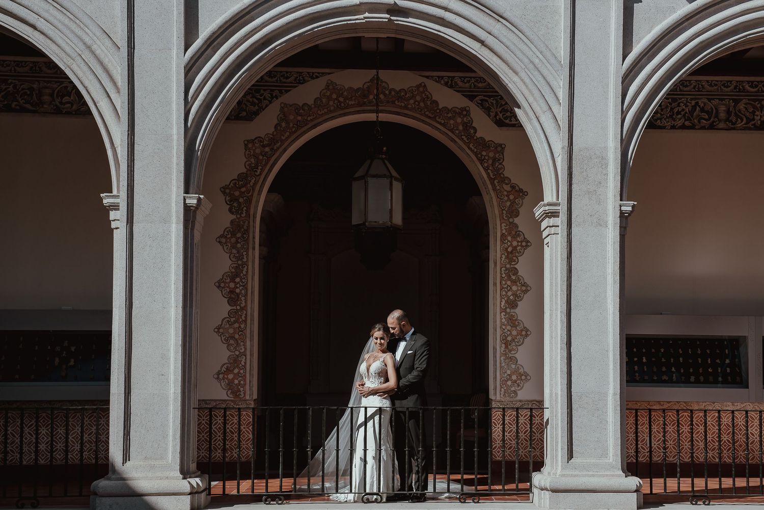 Bride and groom embracing under ornate Spanish colonial arches with decorative lantern at elegant wedding venue.