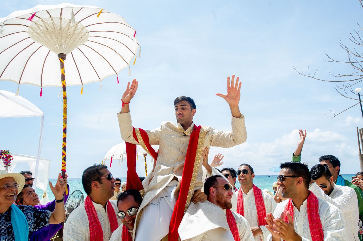 Groom lifted by guests during Indian beach wedding ceremony with white umbrella and red sashes under blue sky.