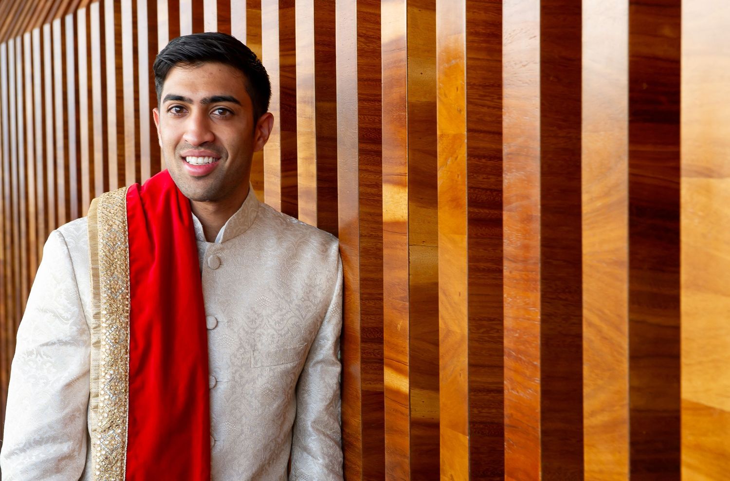 Young man in traditional Indian sherwani with red dupatta smiling against wooden striped wall.