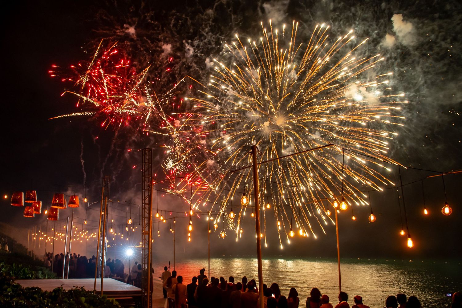 Colorful fireworks exploding over a waterfront crowd at night, illuminating the dark sky with red and gold bursts.
