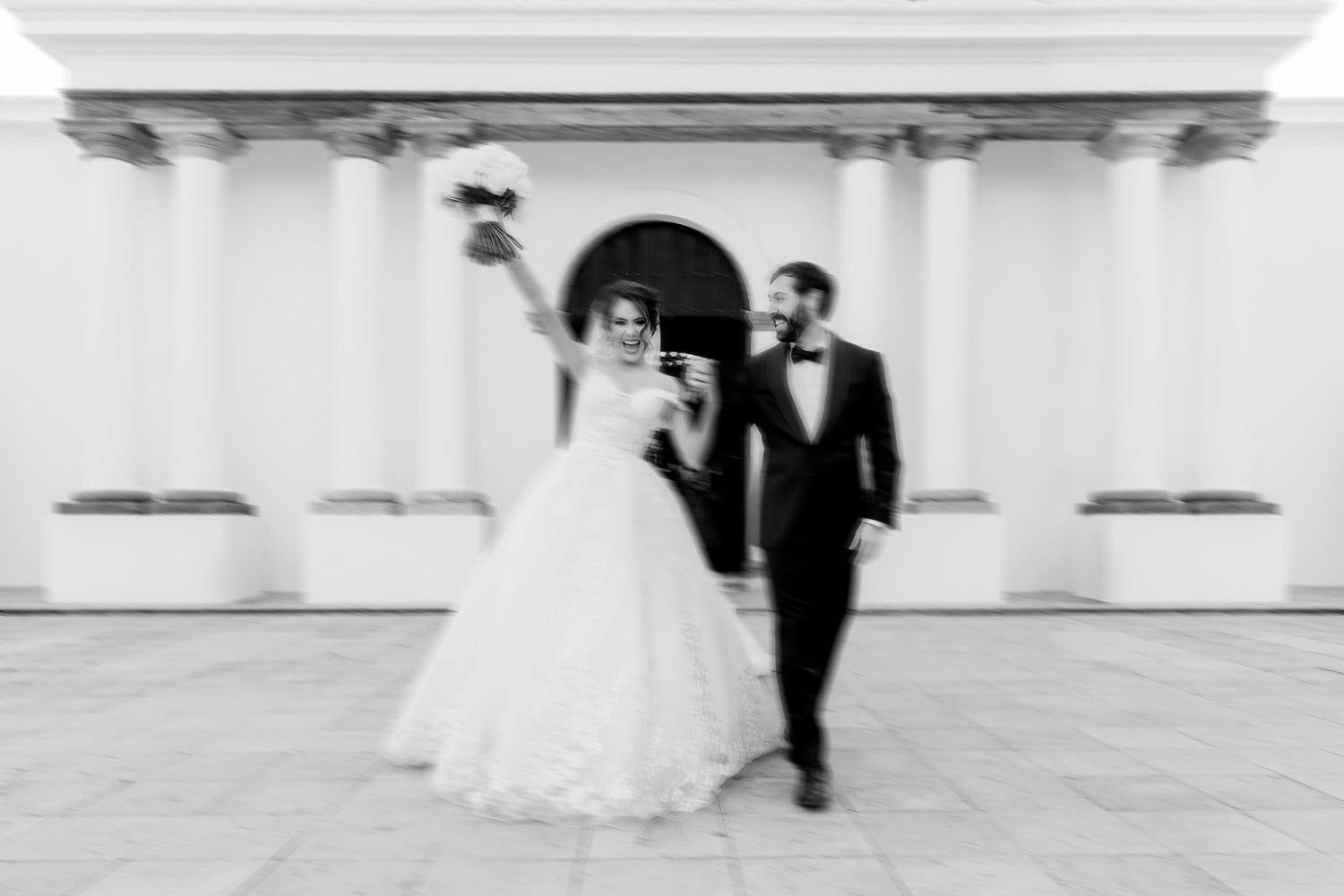 A black and white wedding photo showing a couple dancing in an elegant columned venue.