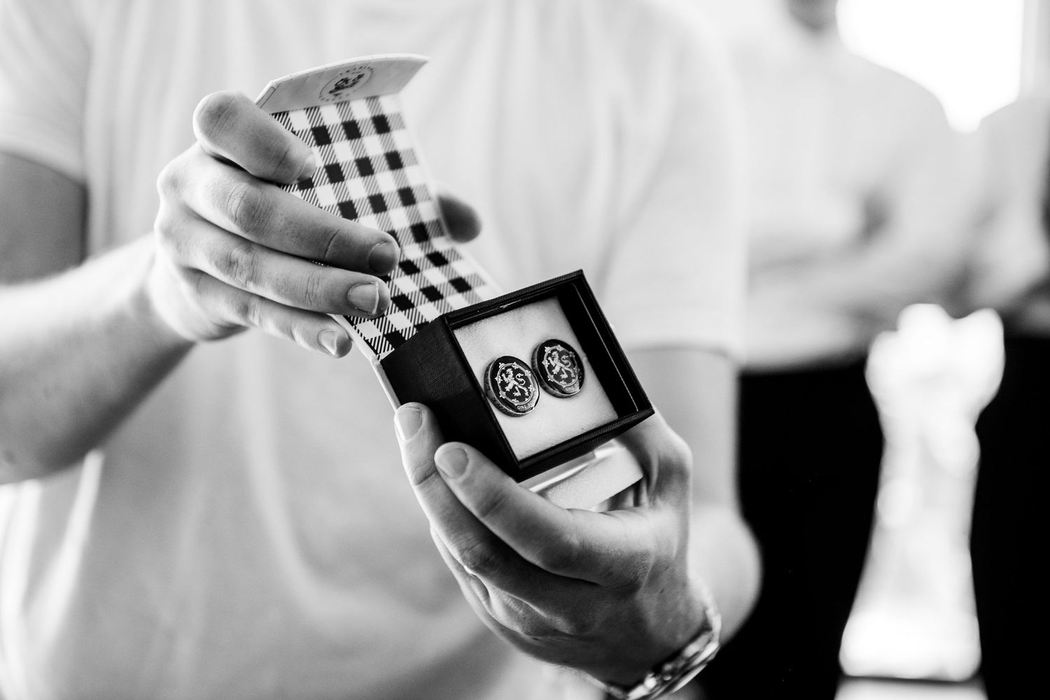 Close-up black and white photo of hands holding cufflinks in a gift box with a checkered tie.